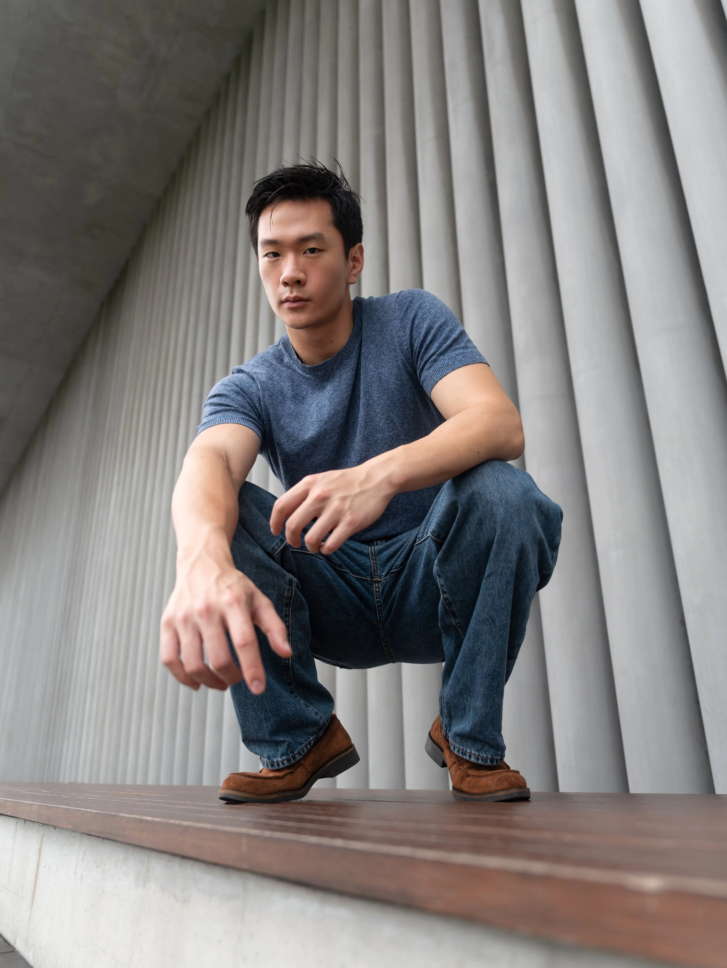 A young man with dark hair, wearing a blue t-shirt, jeans, and brown boots, crouching on a wooden platform against a backdrop of vertical metallic wall panels.