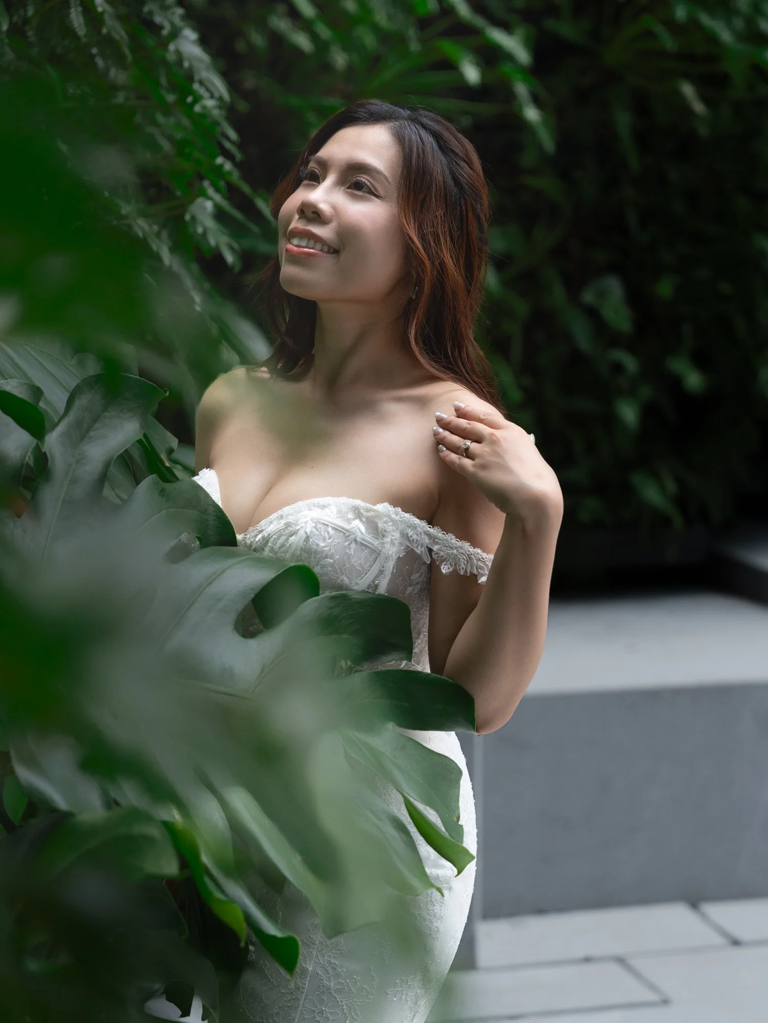 A smiling woman with long brown hair, wearing a white dress, standing outdoors among green foliage.