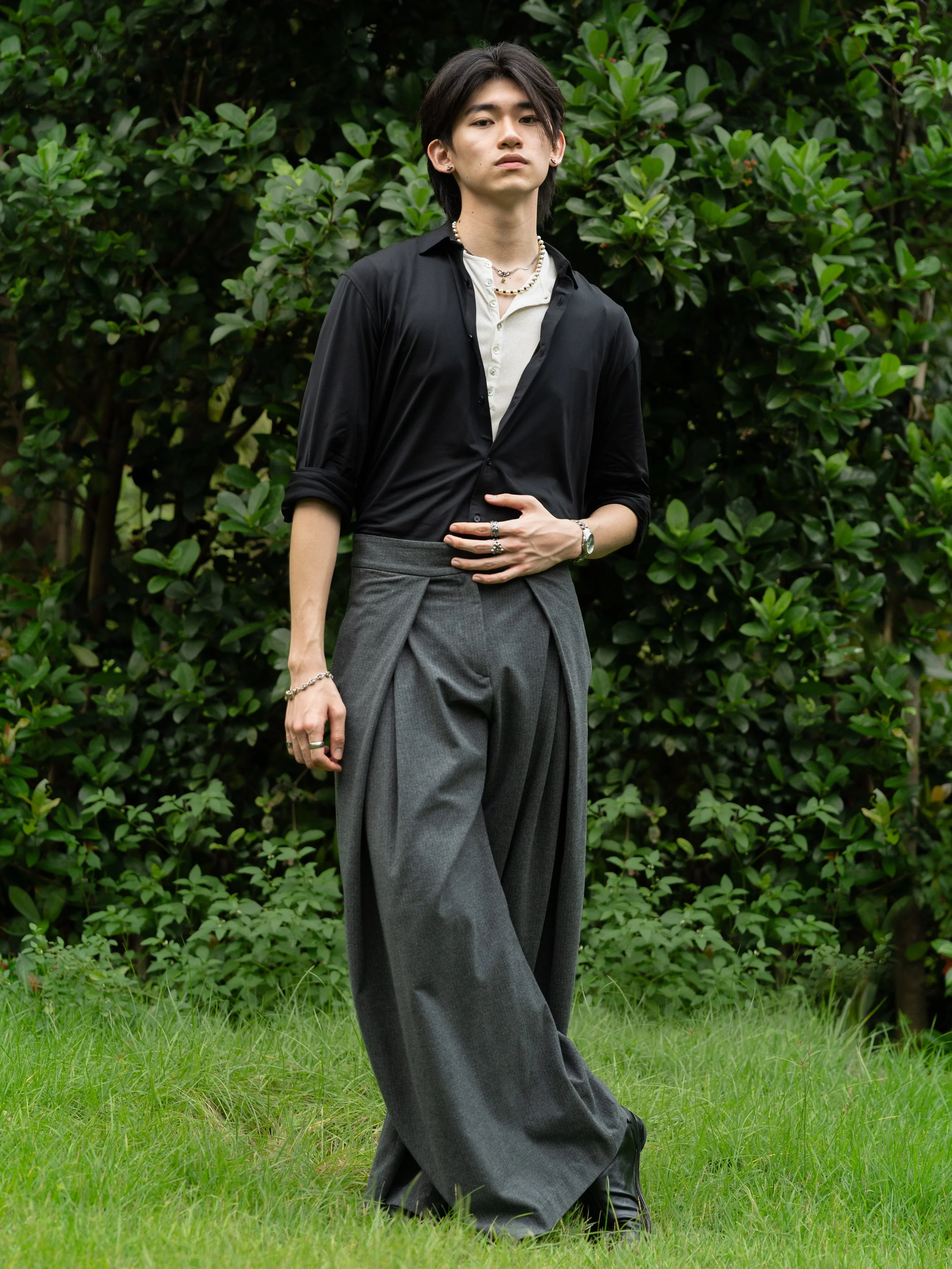 A young man standing outdoors in front of a large green bush, wearing casual stylish clothing including a black shirt, gray wide-leg pants, jewelry, and black shoes.