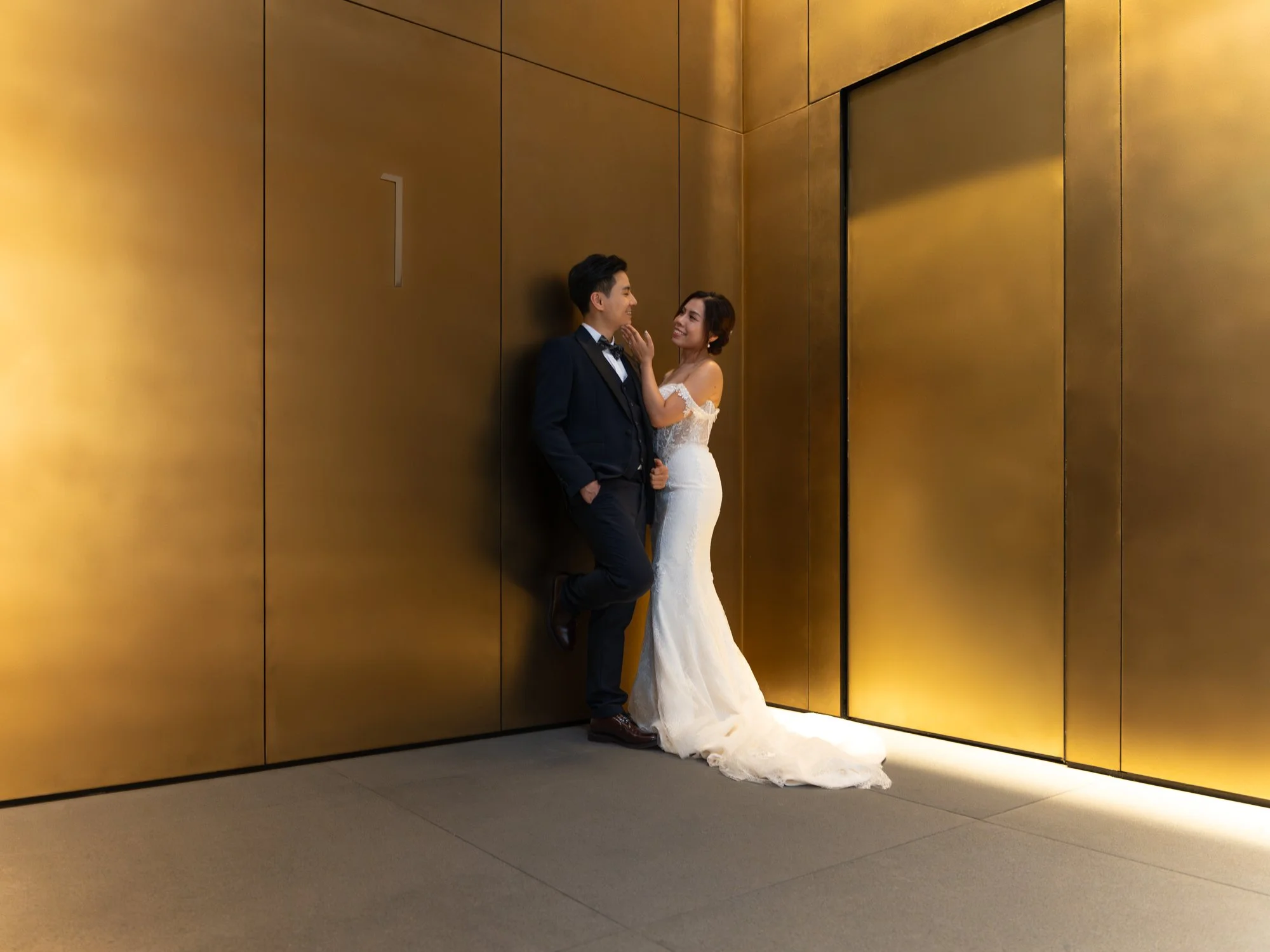 A bride and groom standing close together against a gold-colored wall in an indoor setting, smiling and gazing at each other. The bride is wearing a white lace wedding gown with off-the-shoulder sleeves, and the groom is in a black suit with a bow ti