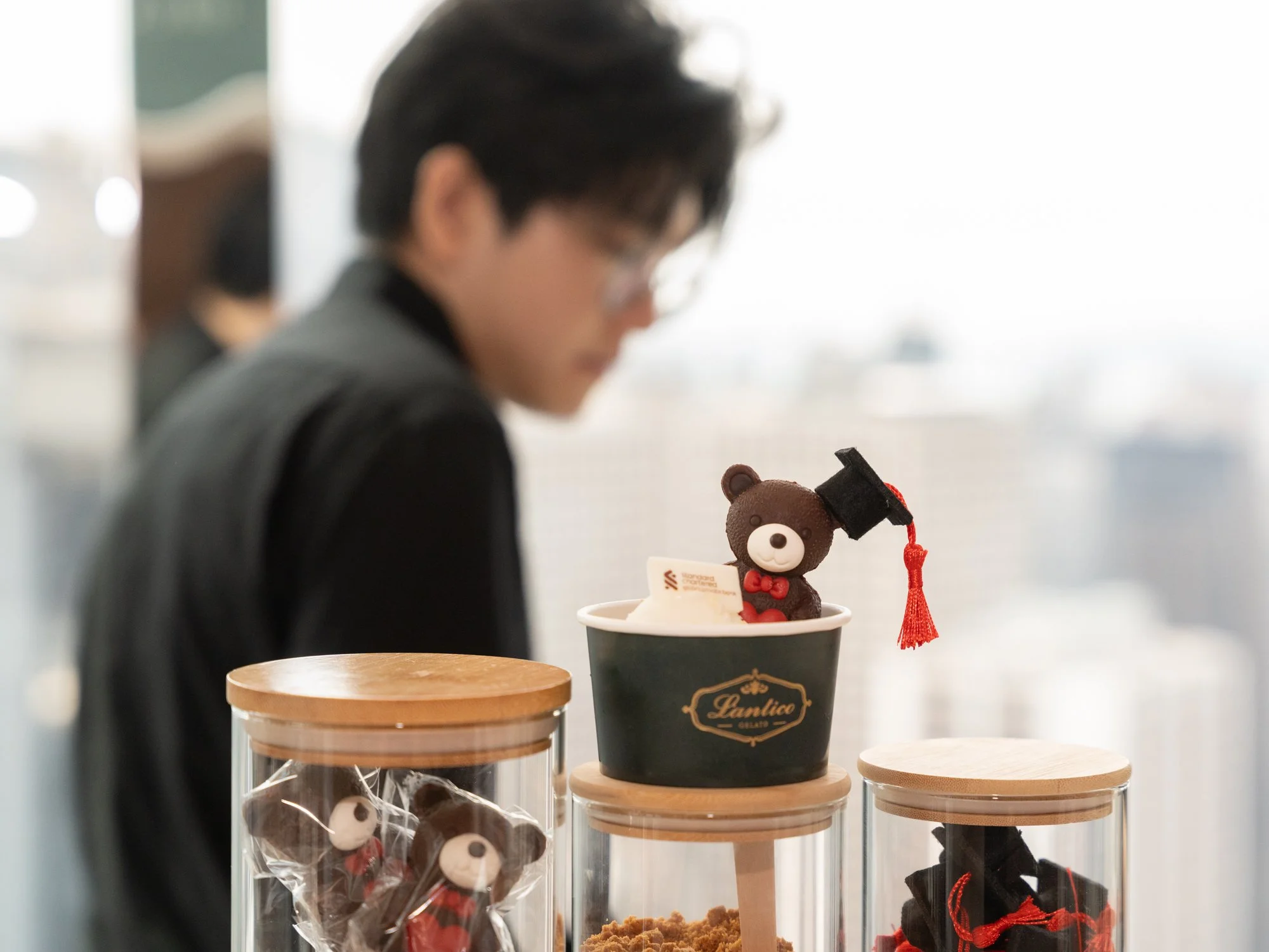 A person with dark hair and glasses looking at their phone, with a small teddy bear wearing a graduation cap and red bow on a surface in front of them. The bear is in a black cup with a golden logo, alongside other containers with baked goods and cho