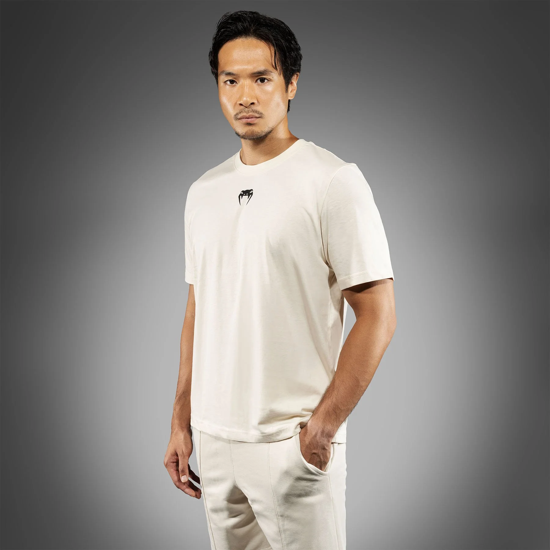 A young man with dark hair, wearing a cream-colored t-shirt with a small black logo on the chest, and matching cream pants, standing against a gray background.