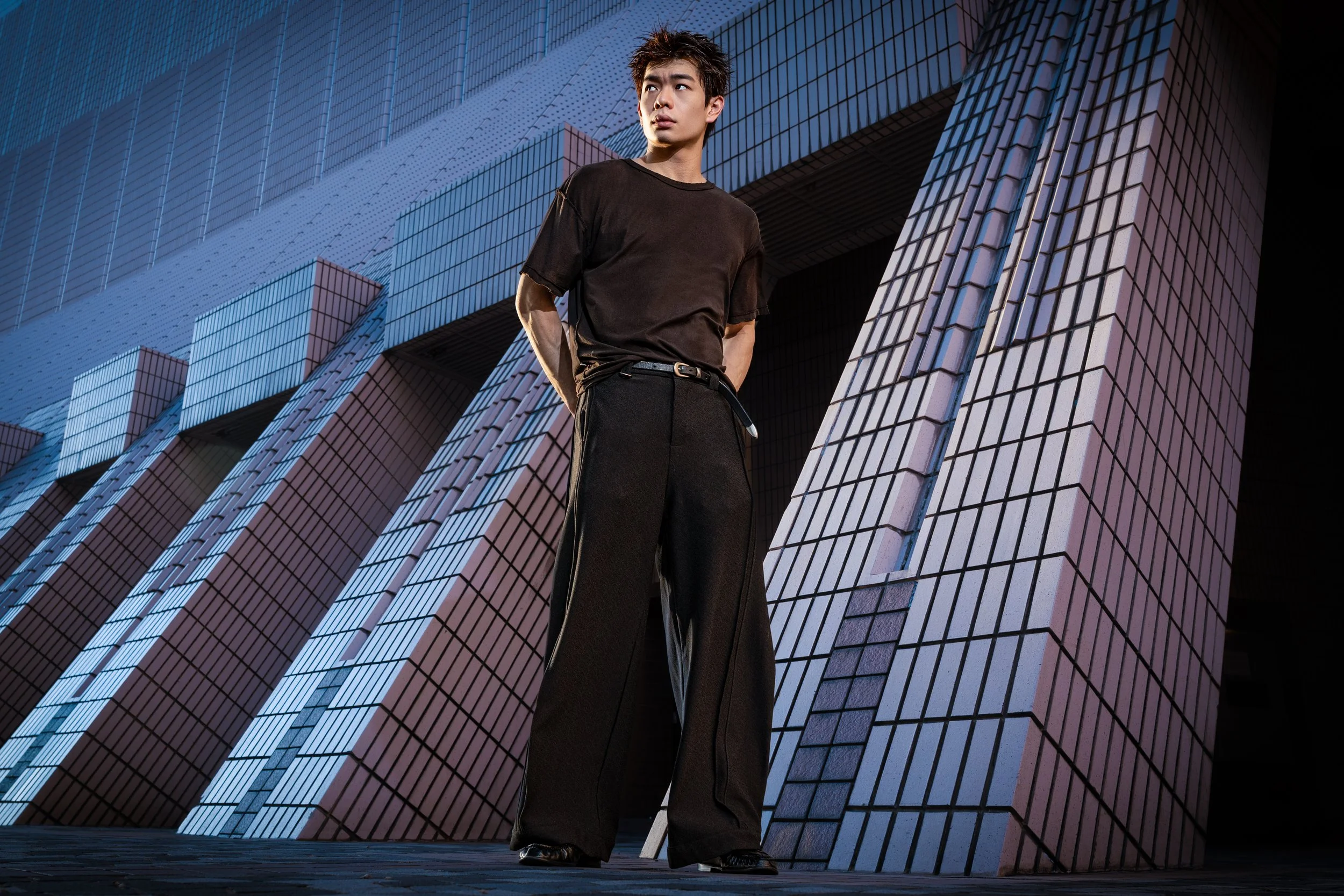 Young man with spiky hair wearing a loose black T-shirt and black pants standing in front of a modern, tilted glass building.