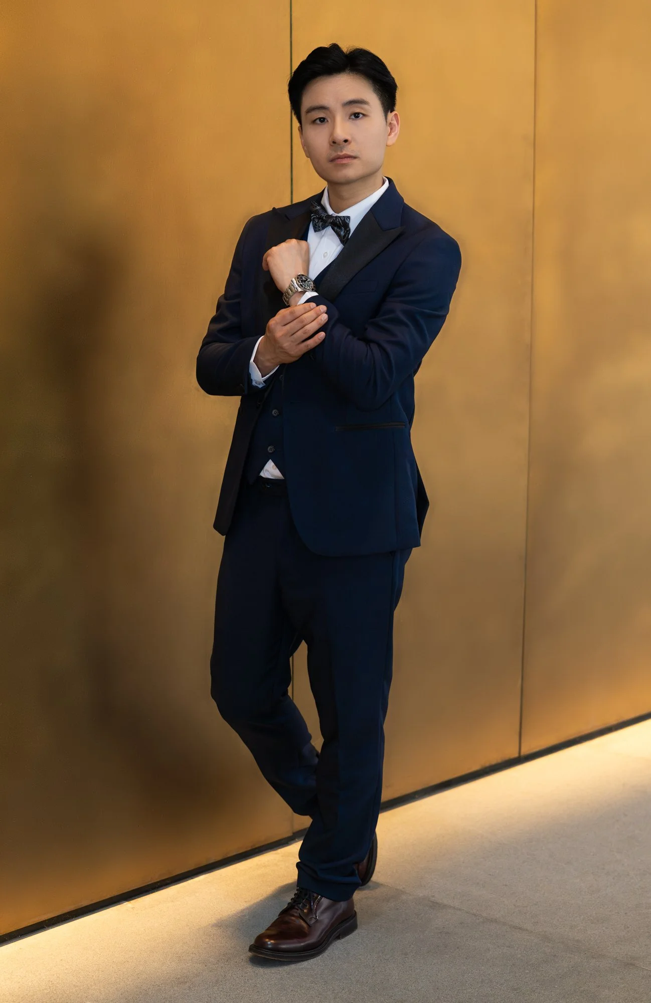 A young man in a navy blue tuxedo with a bow tie and brown dress shoes, standing against a gold wall, looking at the camera, adjusting his wristwatch.