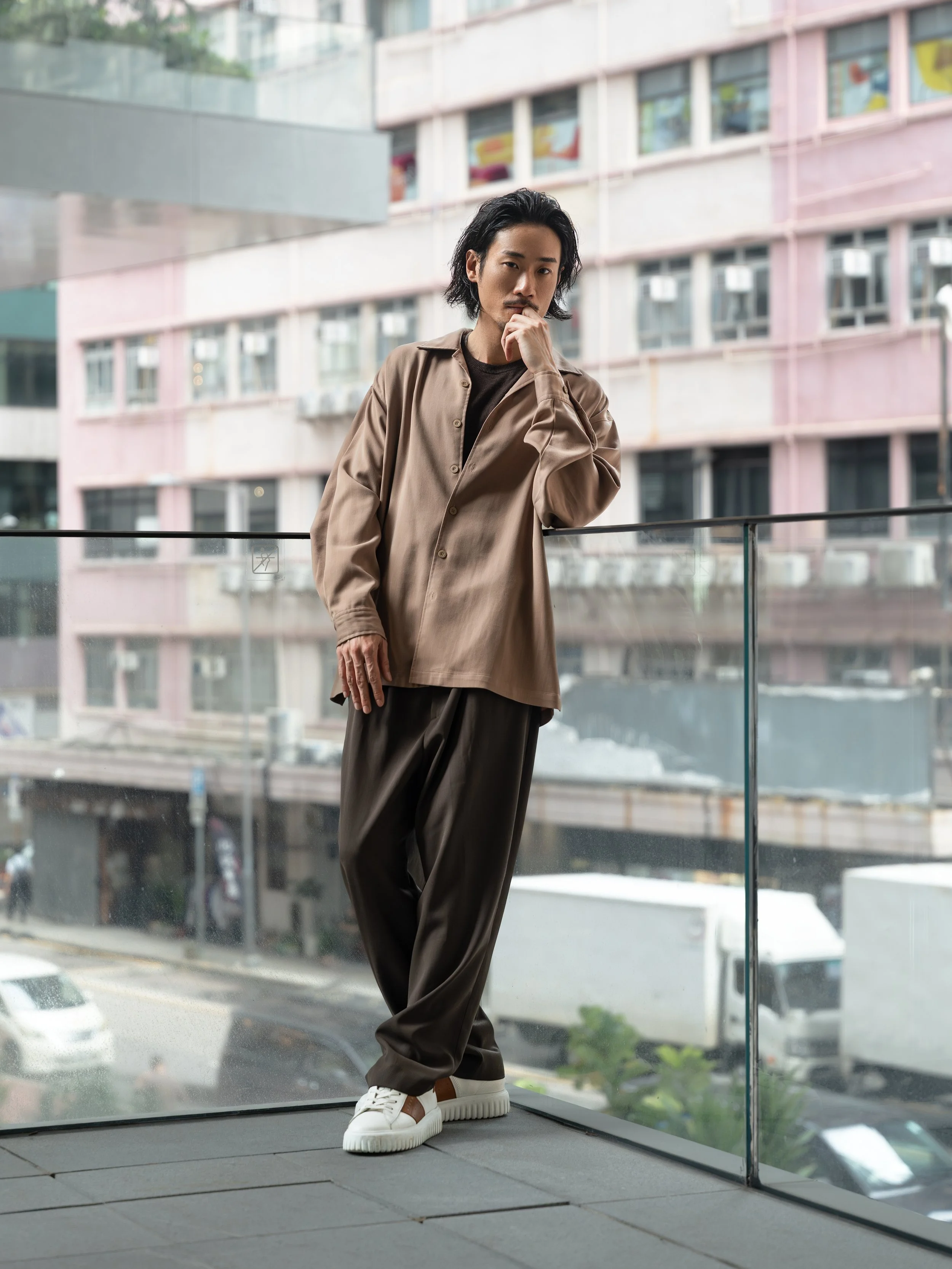 A young man with long black hair, wearing a beige oversized button-up shirt, black pants, and white sneakers, standing by a glass window in an urban setting.