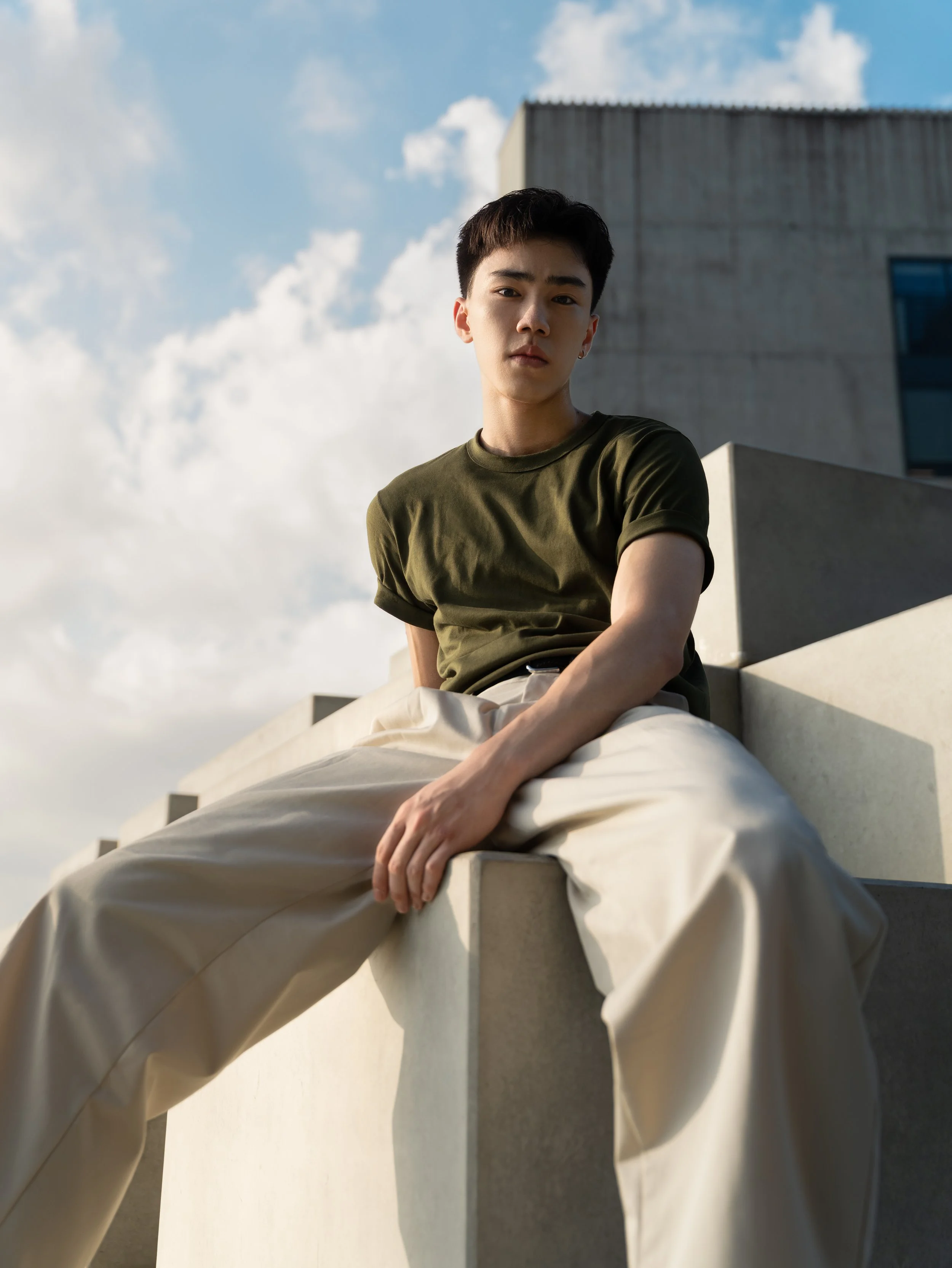 A young man with short dark hair wearing a green T-shirt and beige pants sitting on a modern concrete ledge outdoors, with a blue sky and scattered clouds in the background.
