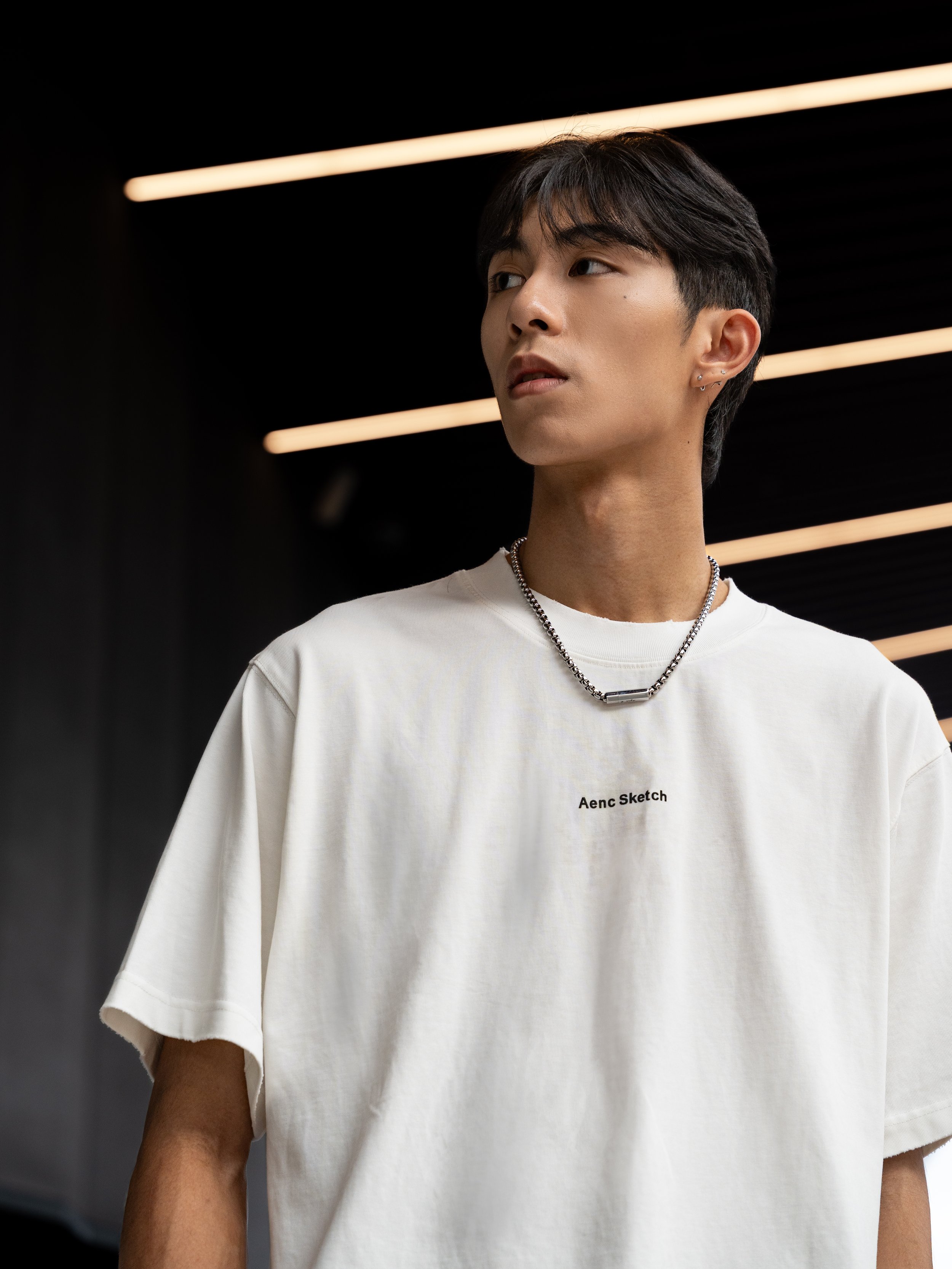A person wearing a white T-shirt with black text that reads 'Aenc Sketch,' silver chain necklace, and earrings, standing in front of a black background with linear lighting.