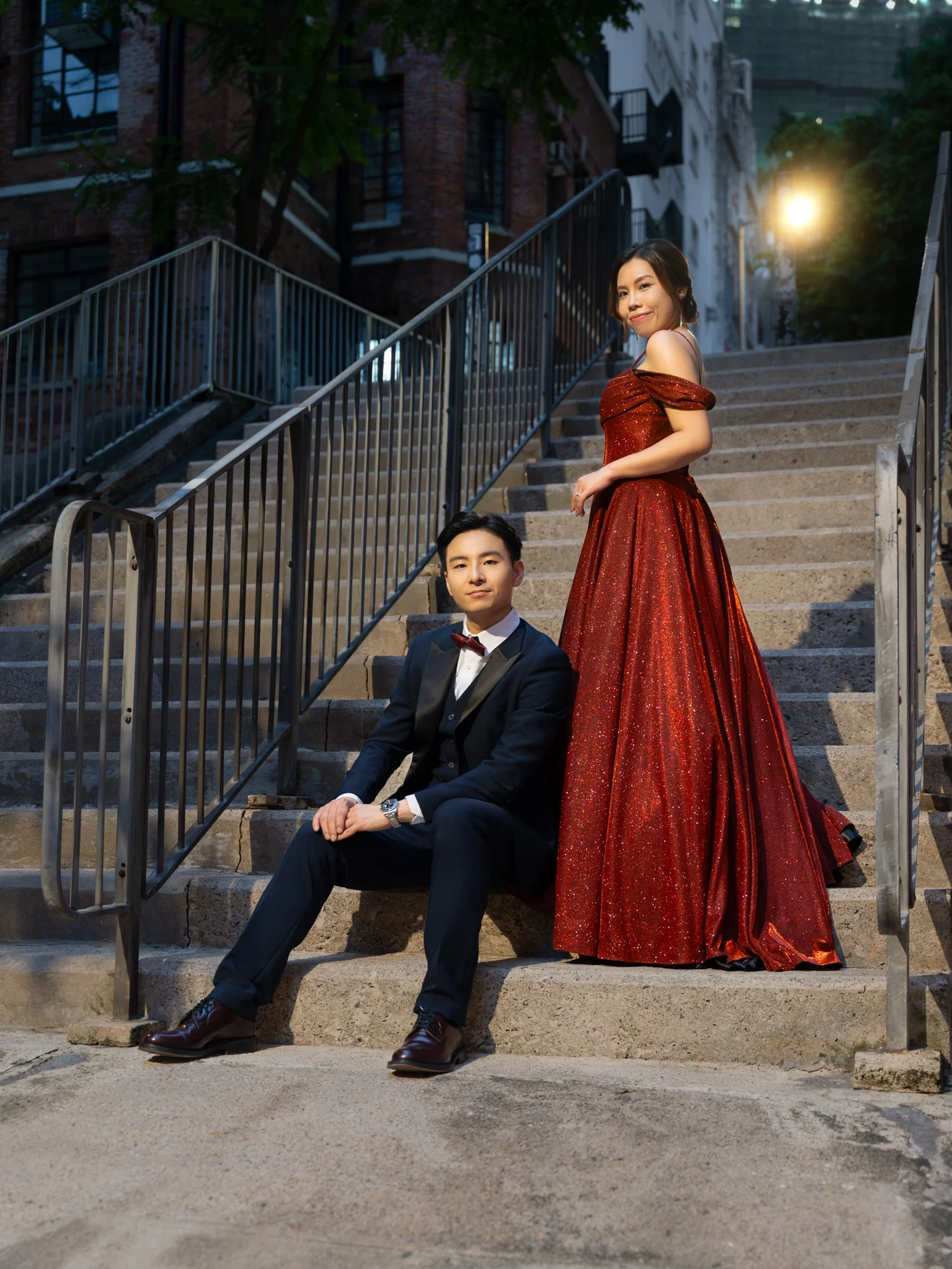 A well-dressed man in a tuxedo with a bow tie sitting on outdoor steps, and a woman in a sparkling red gown standing beside him on stairs, with city buildings and trees in the background