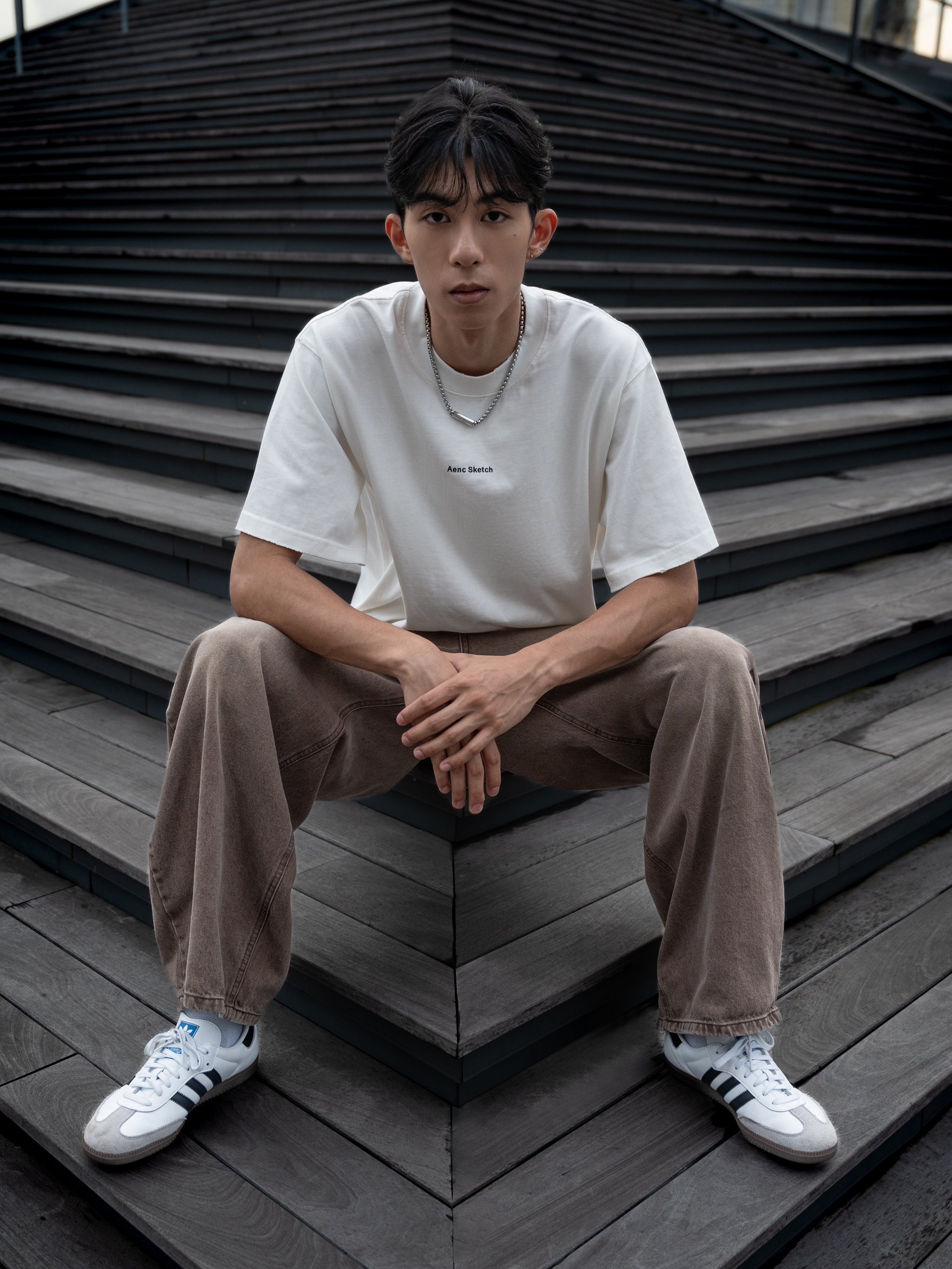 Young man sitting on outdoor wooden steps, wearing a white T-shirt, beige pants, white sneakers, with chains around his neck, in an urban setting.
