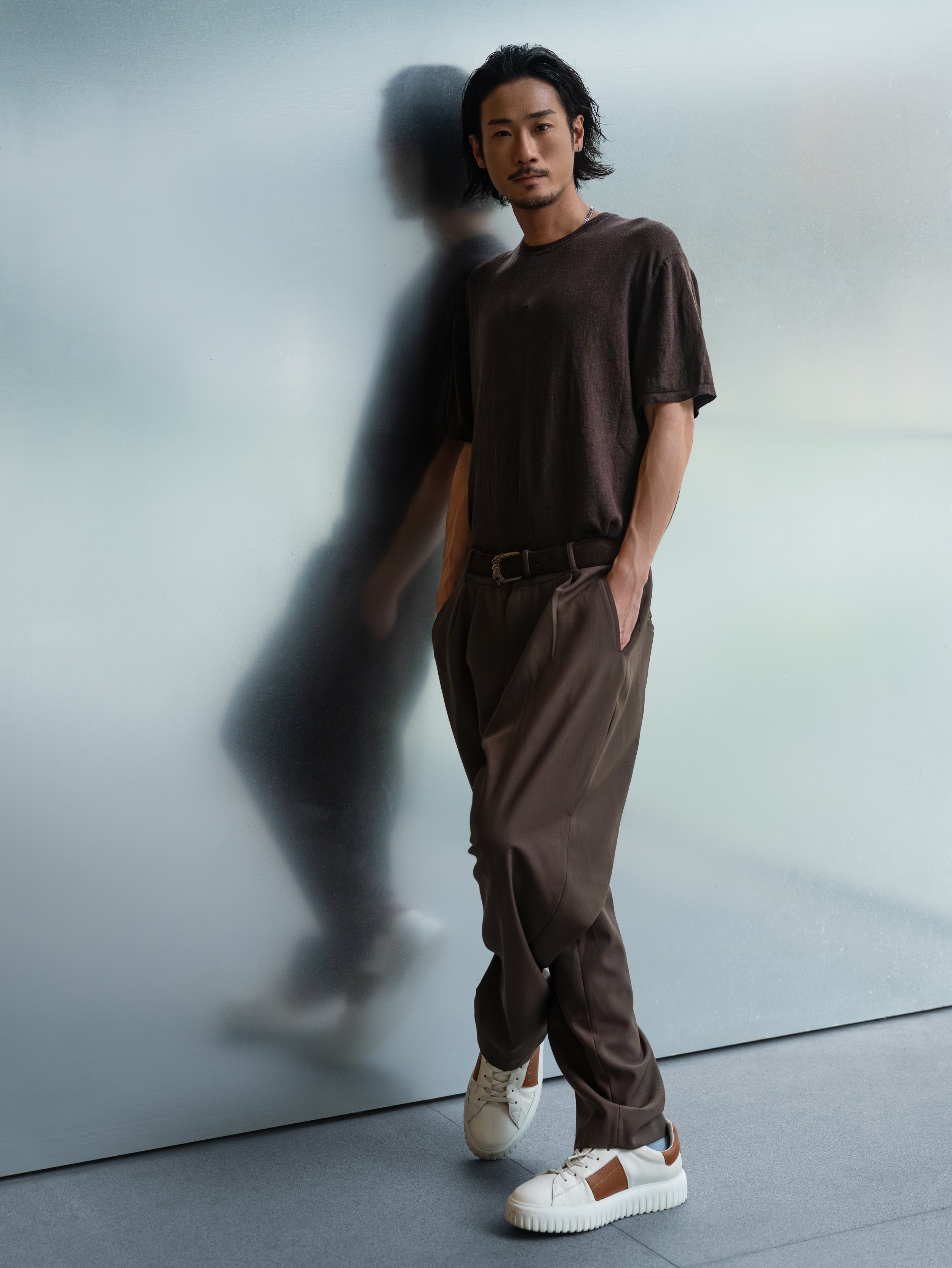 A man with long black hair and a mustache poses against a plain, muted wall. He is wearing a dark brown t-shirt, brown pants, and white sneakers with brown accents.