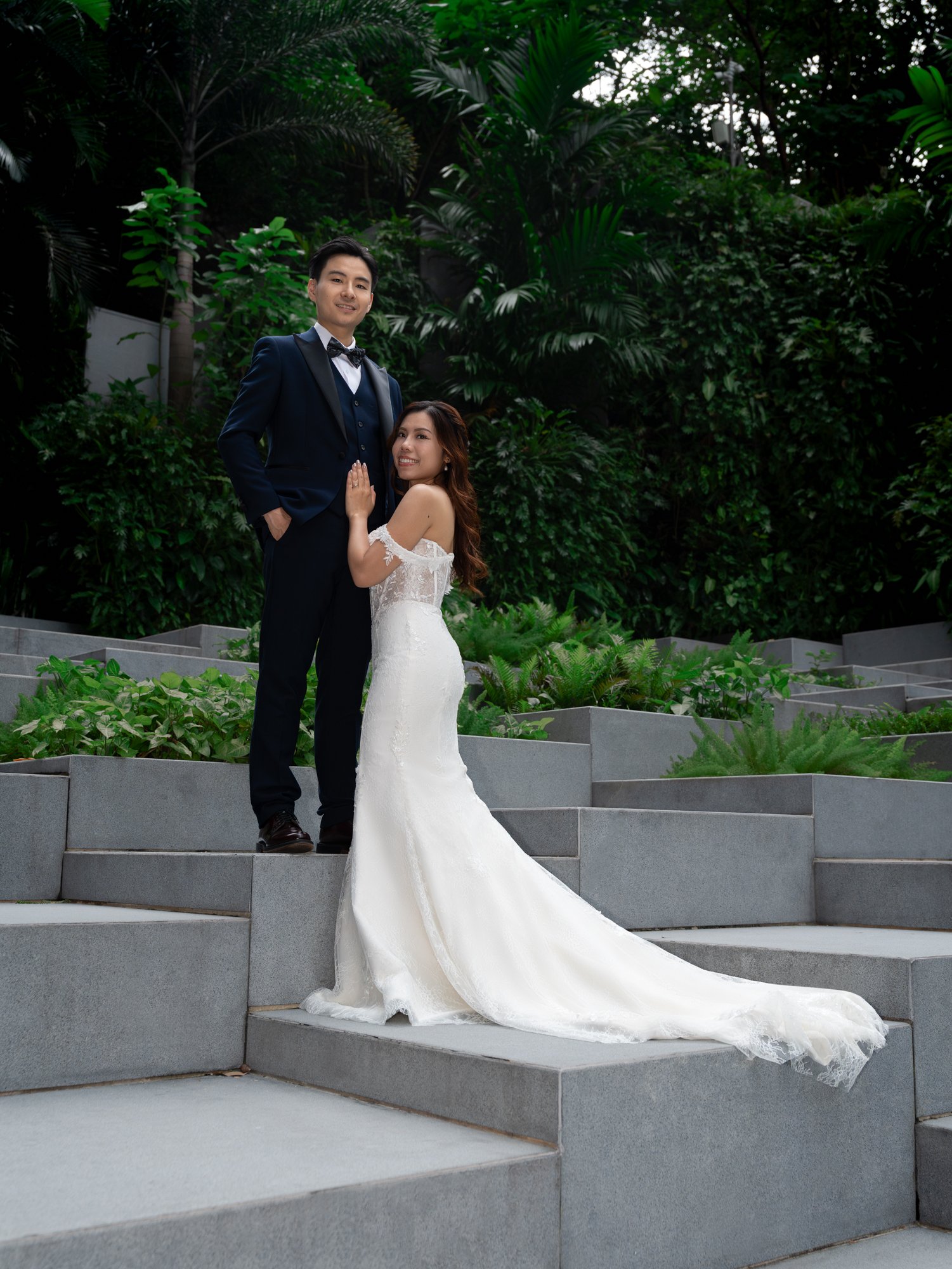 A newlywed couple stands on gray stairs surrounded by lush green plants and trees. The groom is dressed in a navy blue tuxedo with a black bow tie, and the bride wears a white off-shoulder wedding gown with a long train. They are smiling and looking 