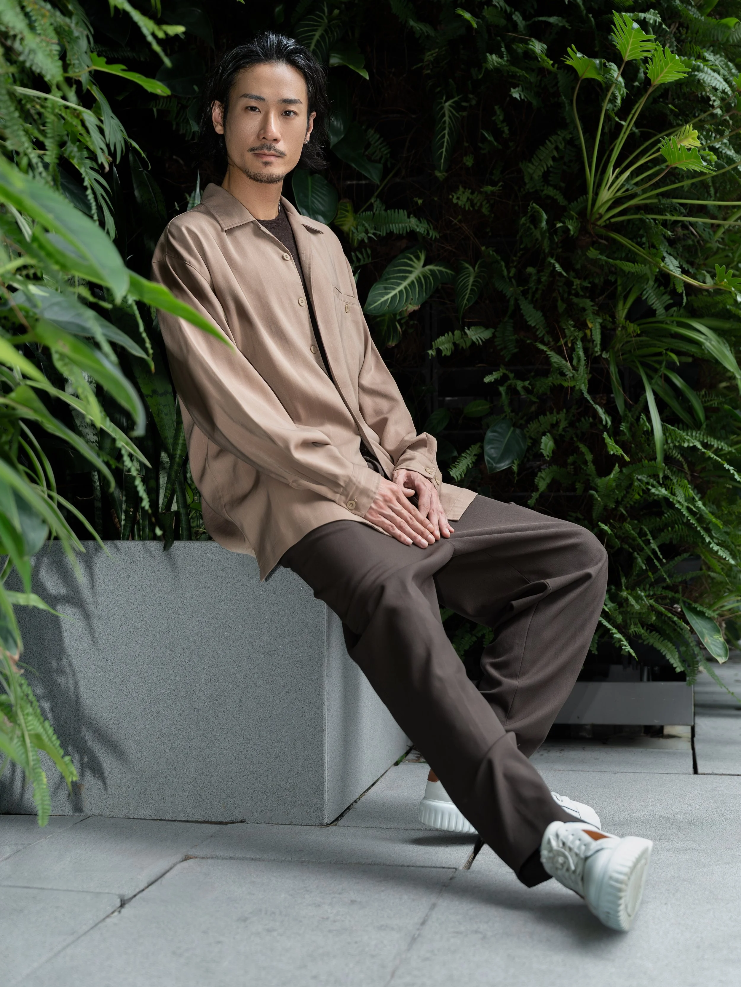 A man sitting on a bench in an outdoor garden surrounded by lush green plants, wearing a beige jacket, dark pants, and white sneakers.