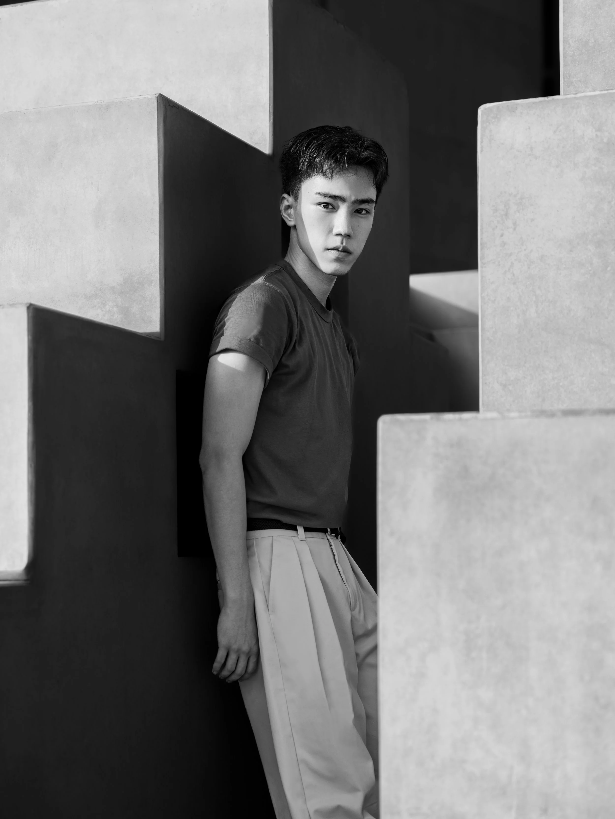 Black and white photo of a young man posing between geometric concrete walls, looking at the camera.