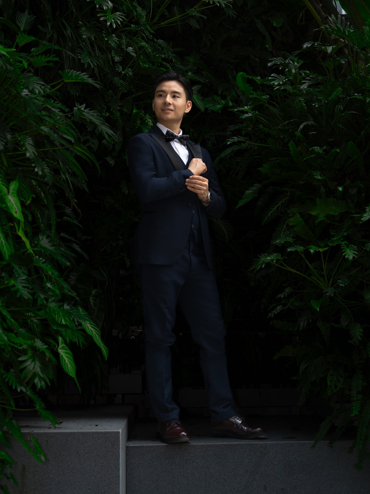 A man in a dark suit with a bow tie stands on steps in front of lush green tropical plants, looking away from the camera.
