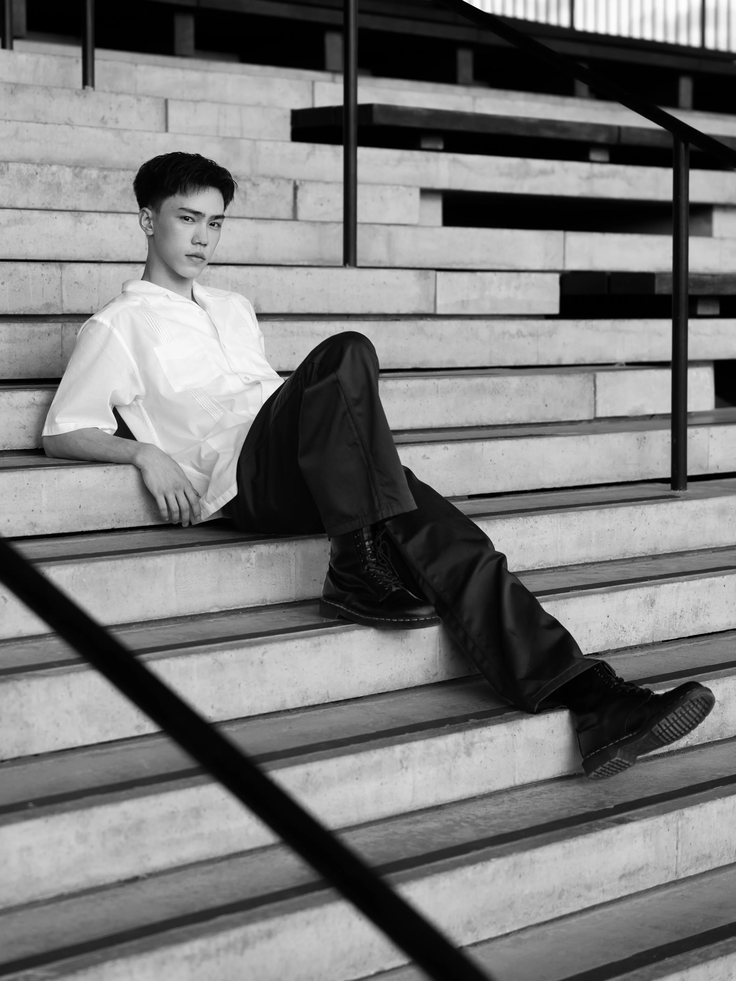 A young person with short dark hair sitting on concrete stairs in an urban setting, wearing a white shirt and dark pants