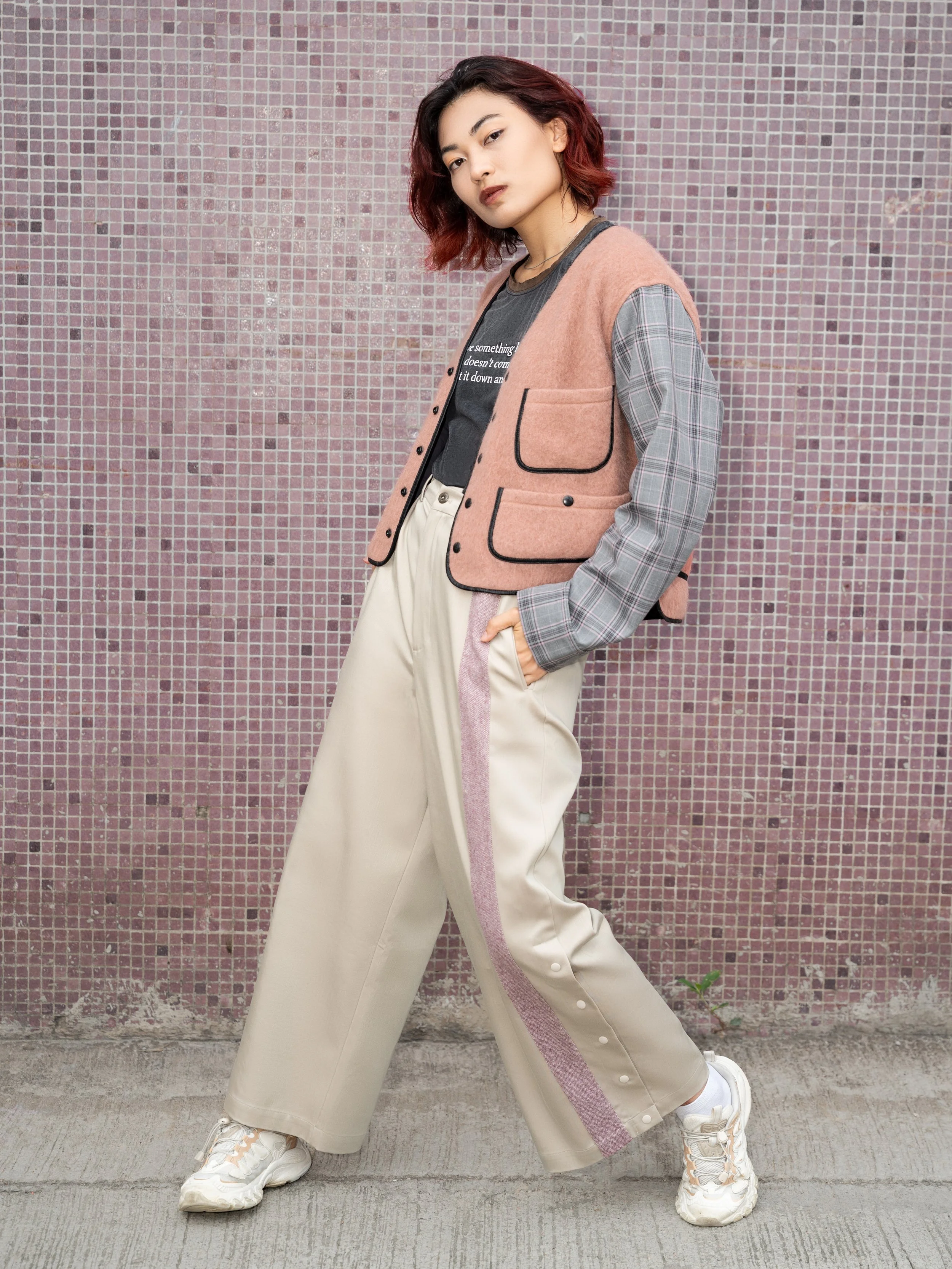 Person with short wavy hair wearing a pink and gray jacket, wide-legged beige pants with pink stripe, and white sneakers, standing against a pink mosaic wall.
