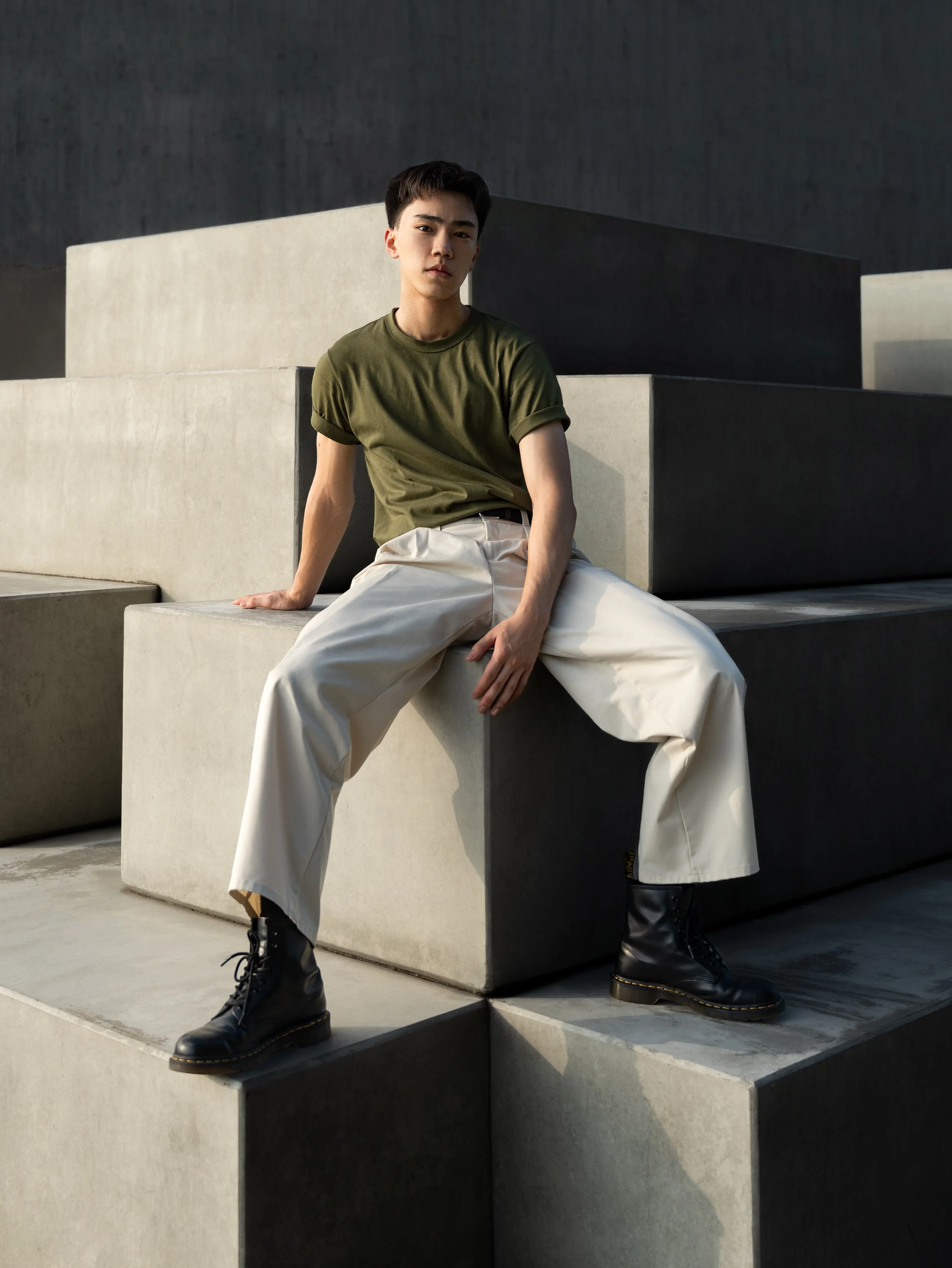 A young man with dark hair sitting on concrete steps, wearing a green t-shirt, wide-leg beige pants, and black combat boots against a minimalist concrete background.
