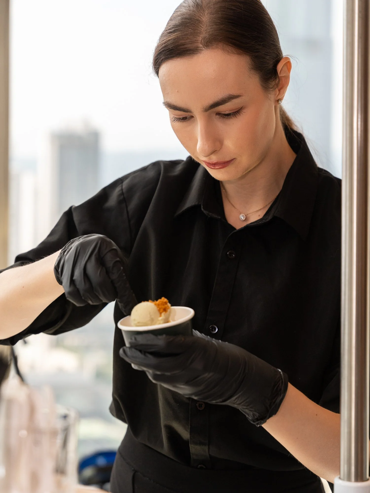 A woman wearing a black shirt and gloves is serving ice cream into a cone, with a blurred background and a bright outdoor setting.