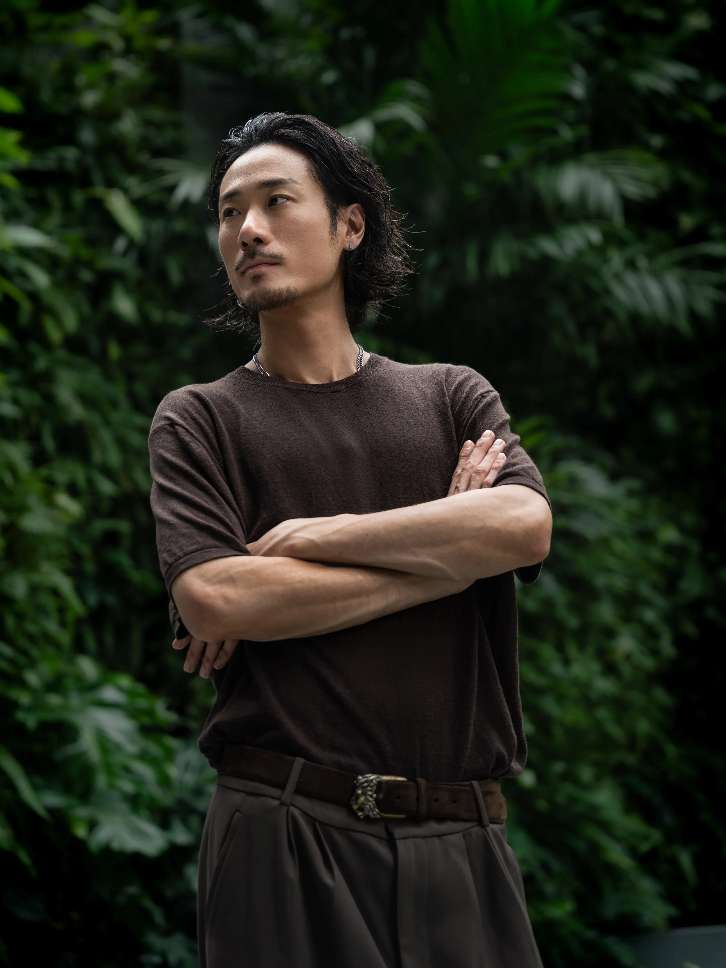 A man with dark hair, a goatee, and earrings, wearing a brown shirt and dark pants, standing with crossed arms in front of lush green foliage.