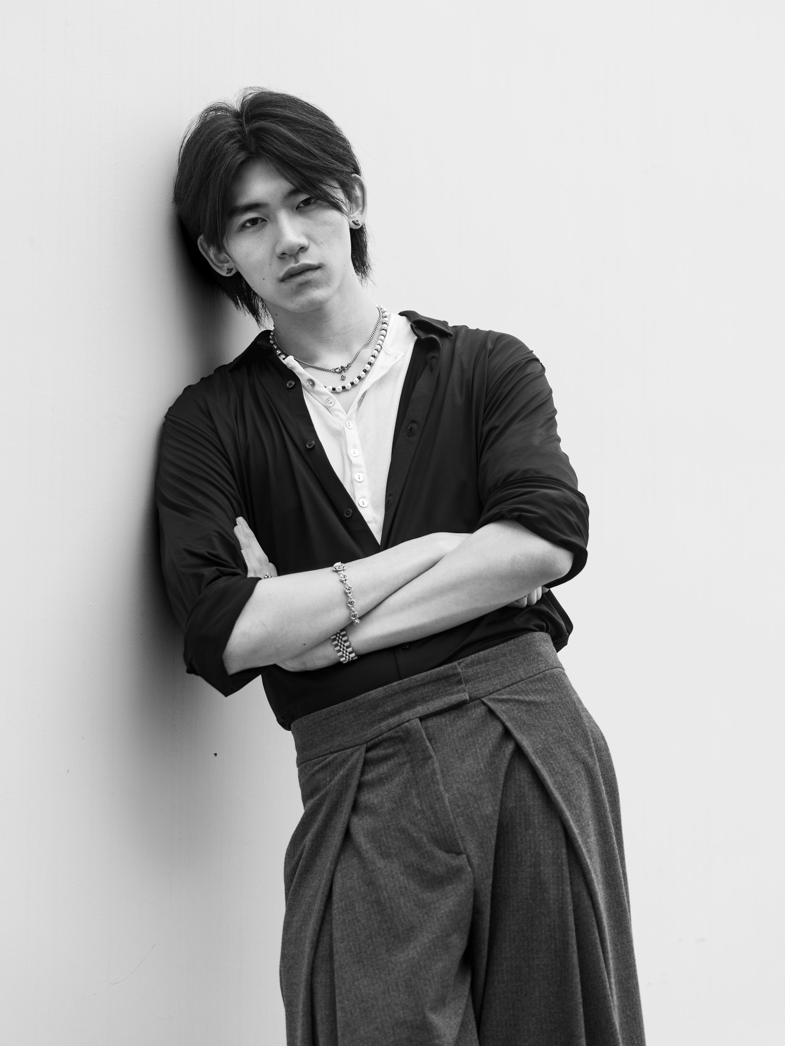 Black and white photo of a young man with medium-length hair, wearing a white shirt, a necklace, earrings, and a dark shirt, leaning against a plain wall with his arms crossed.