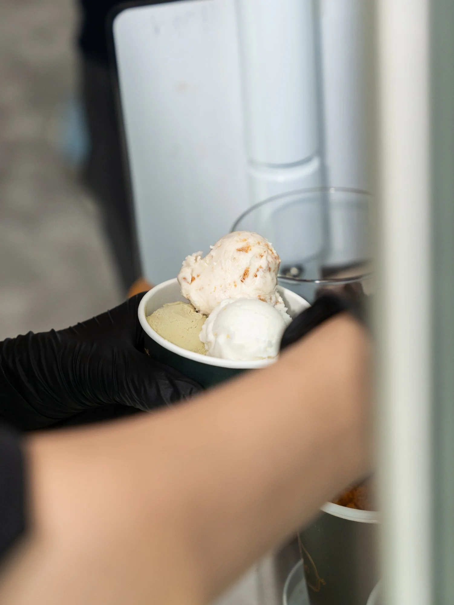 Close-up of a person wearing black gloves holding a bowl of three scoops of ice cream, with an ice cream freezer in the background.