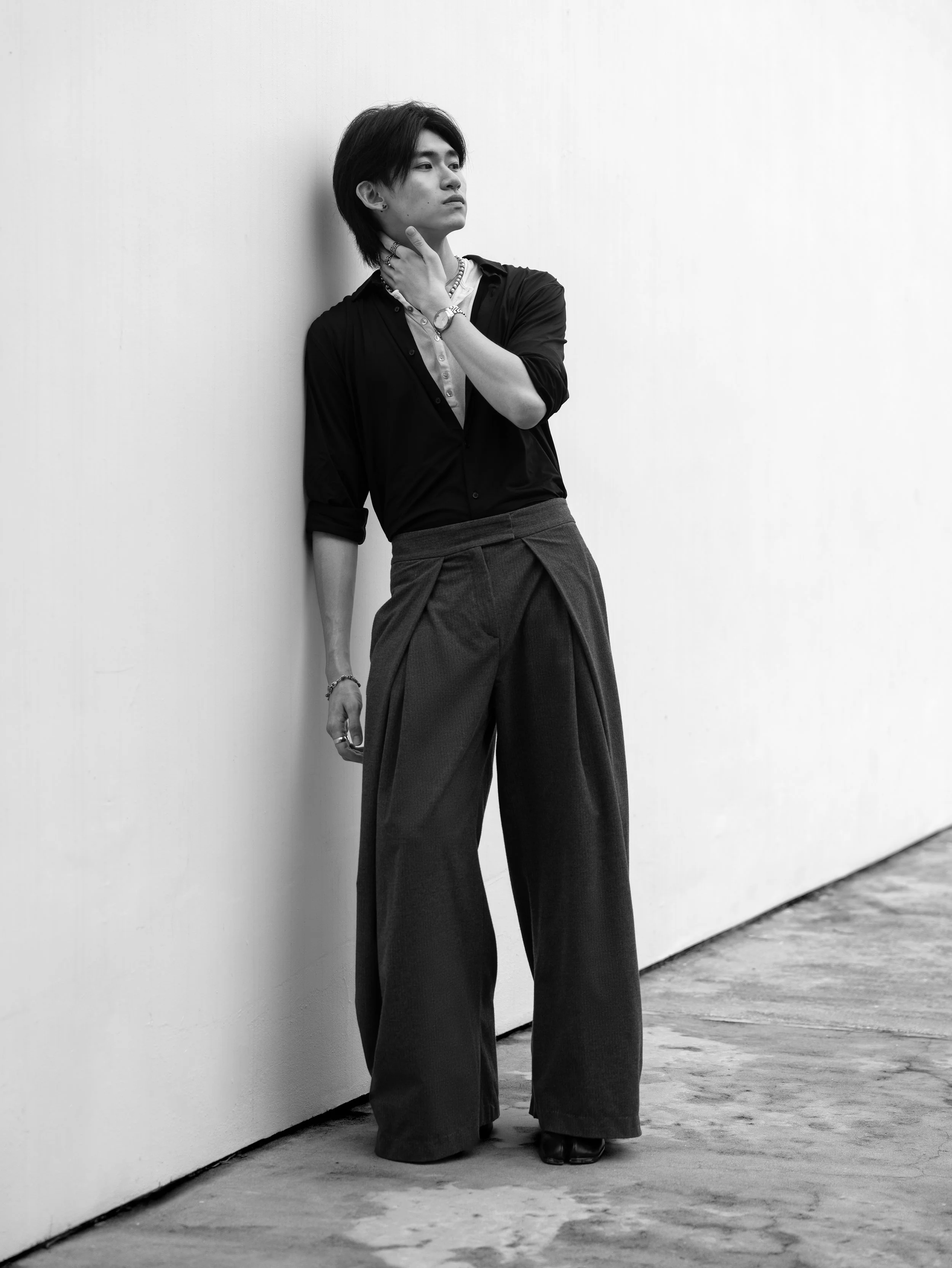 Black and white photo of a woman with short hair, leaning against a plain wall with one hand on her neck and the other hand near her side. She is wearing wide-legged pants, a blouse, and jewelry.
