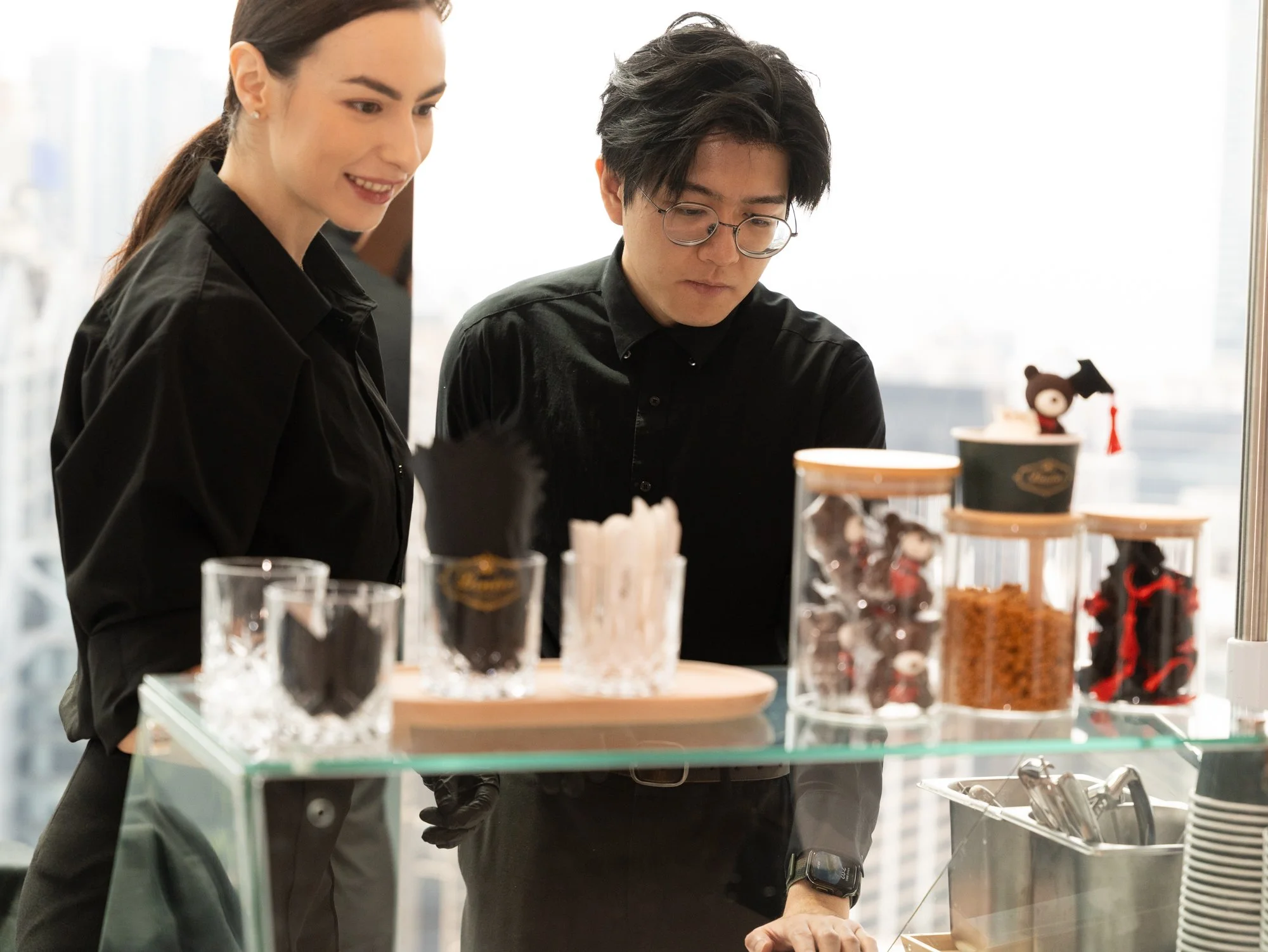 Two people, a woman with dark hair and a man with glasses, are looking at and possibly selecting desserts or snacks displayed on a glass counter. The desserts include various chocolates and decorated treats. The scene is indoors with large windows an