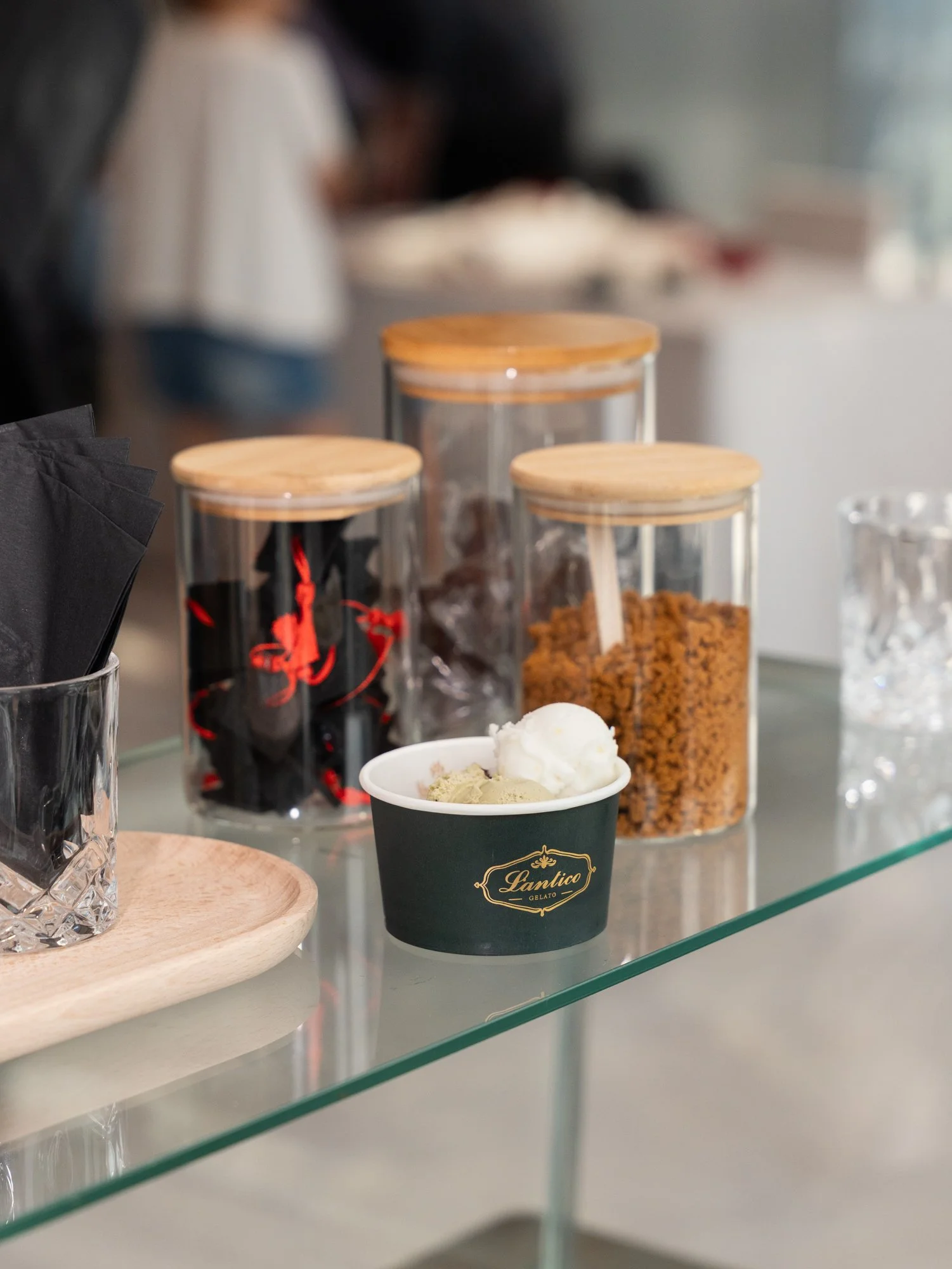 Containers of various flavors of gelato or ice cream on a glass display counter, with one cup of gelato in front labeled "Lantico Gelato".