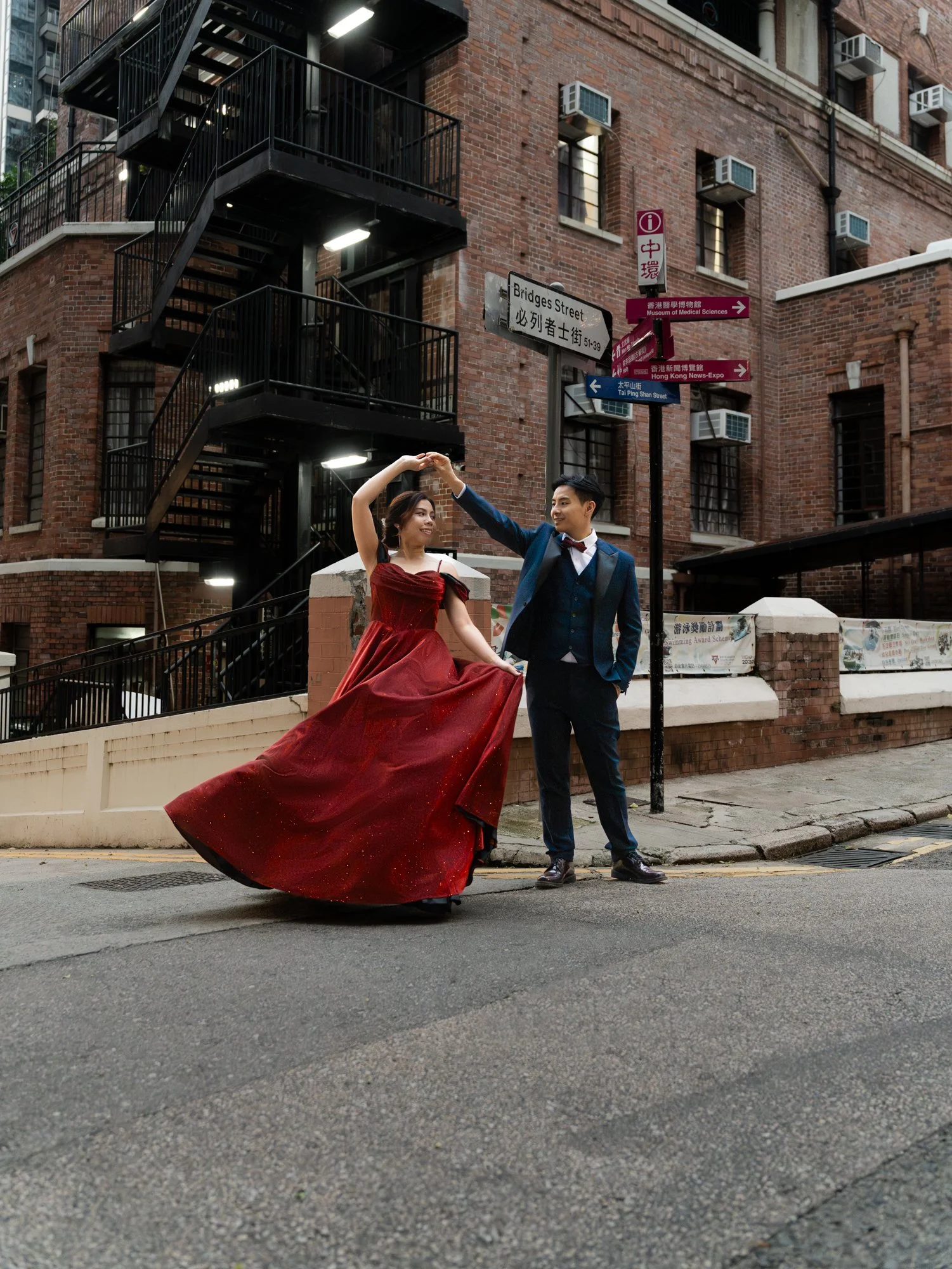 A couple dancing on a city street in front of a brick building, with street signs visible, including one for Bridges Street and another pointing to the Museum of Medical Sciences. The woman is wearing a red dress, and the man is in a suit with a bow 