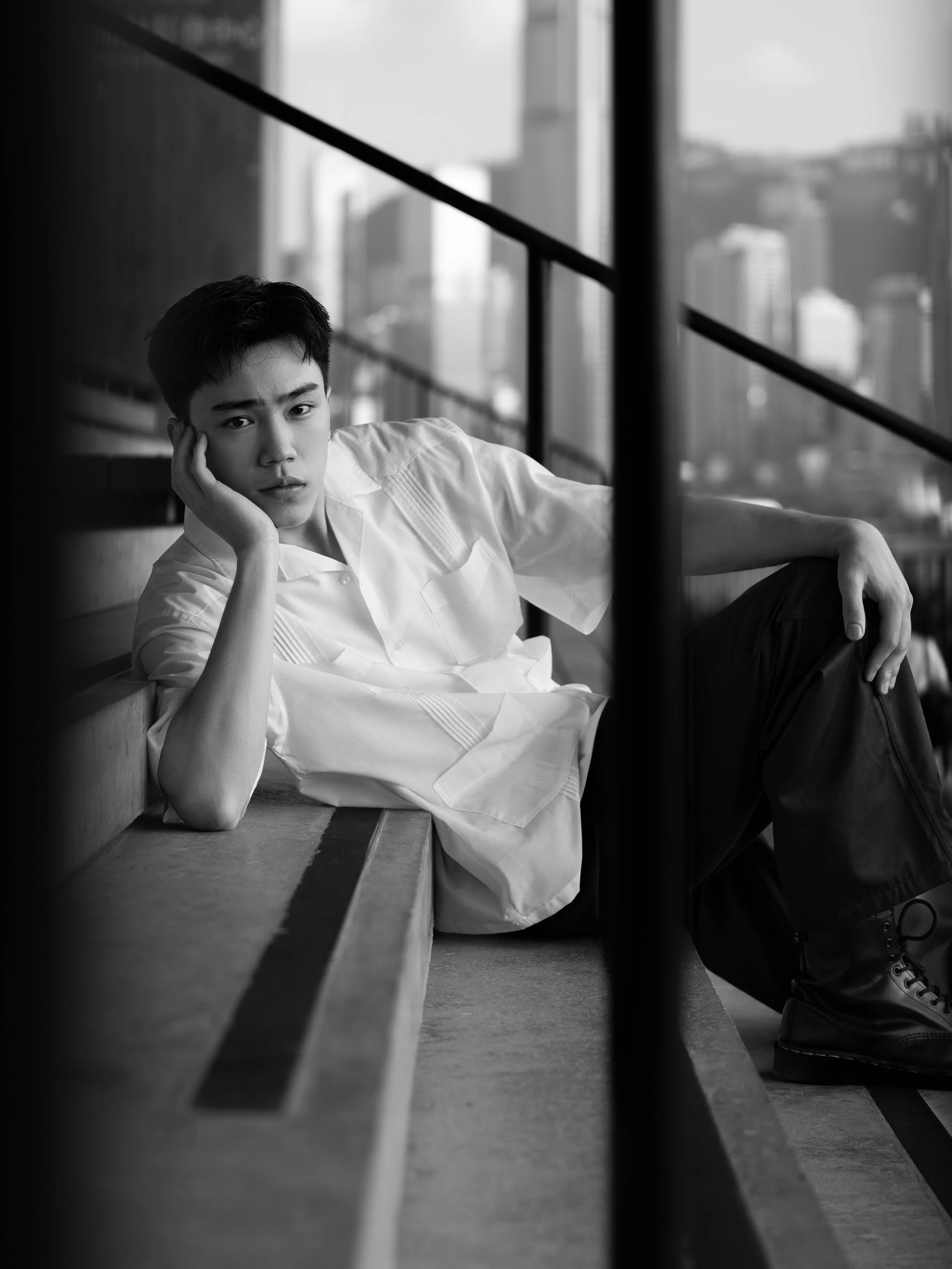 A black-and-white photo of a young man with dark hair, lying on a bench, resting his head on his hand. He is wearing a white shirt, and there is a cityscape with tall buildings visible through the window behind him.