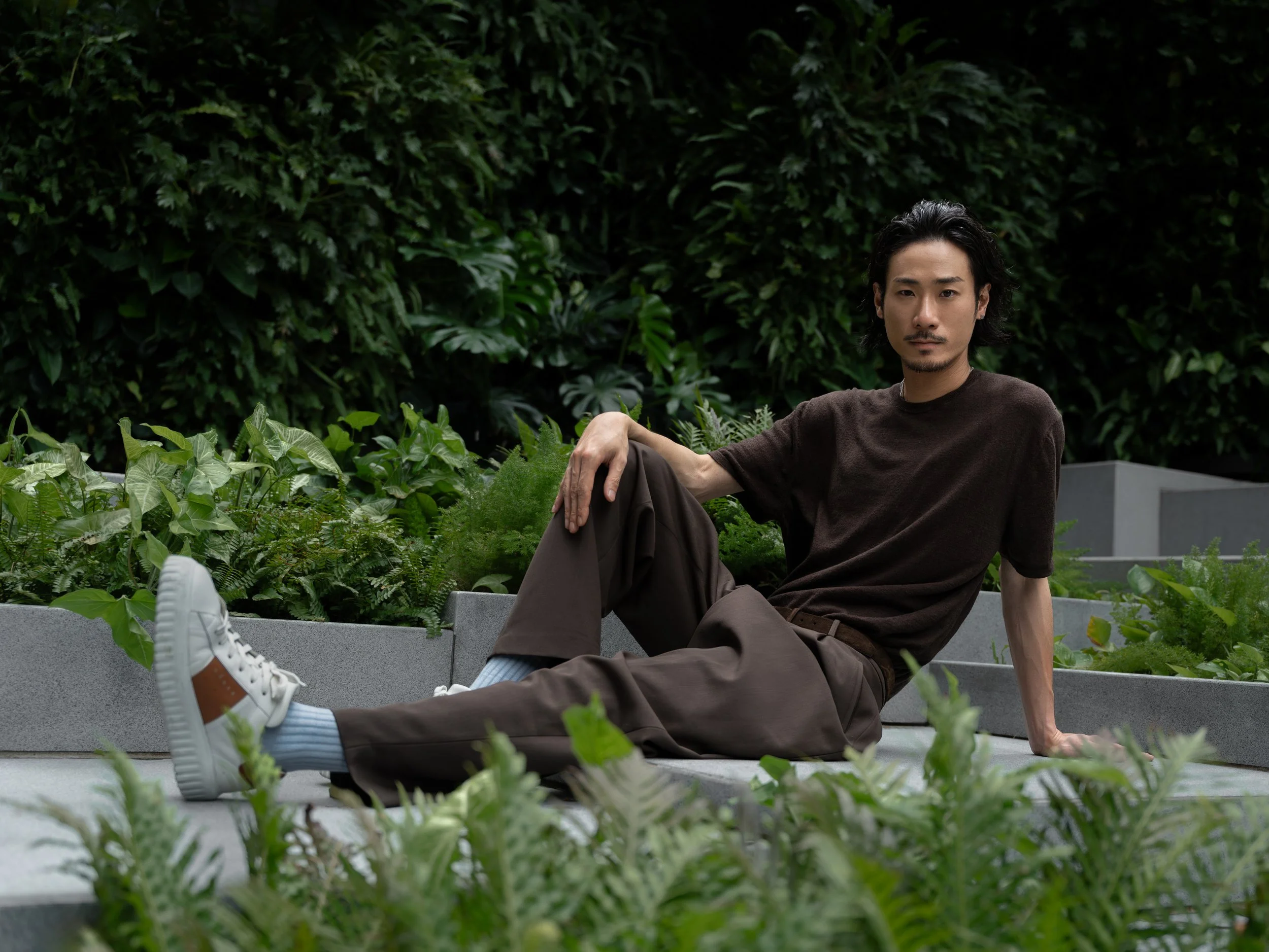 A man sitting outdoors on a concrete surface, surrounded by lush green plants, with a large green leafy background. He is wearing a dark t-shirt, brown pants, and white sneakers, with one arm resting on his knee and the other supporting him on the gr