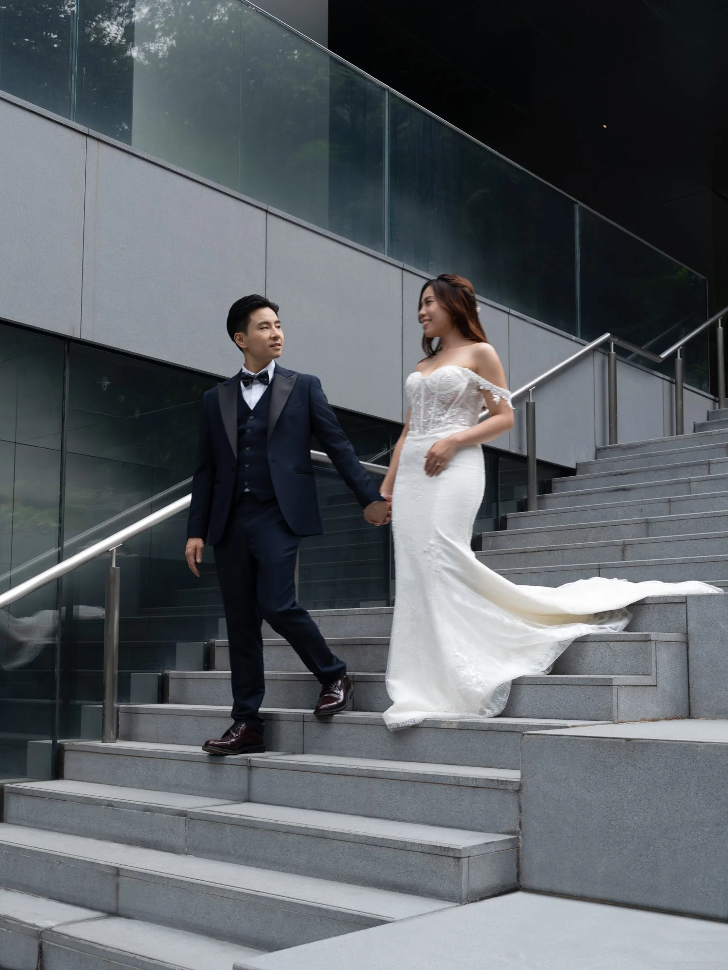 A newlywed couple dressed in wedding attire holding hands and walking down outdoor stairs in an urban setting with modern glass and concrete architecture.