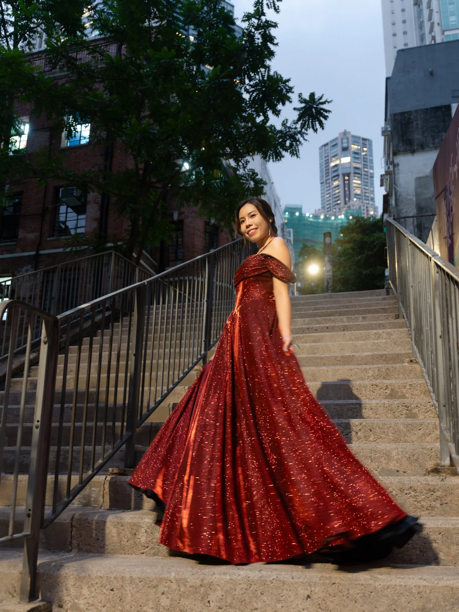 A woman in a red, glittery evening gown stands on outdoor stairs in an urban area at dusk, smiling at the camera with city buildings in the background.