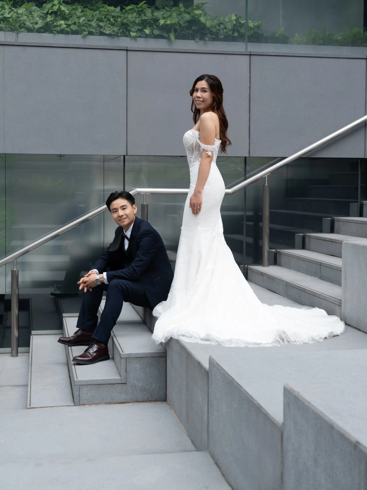 A couple, a woman in a wedding dress and a man in a suit, on outdoor stairs with modern glass and concrete architecture.