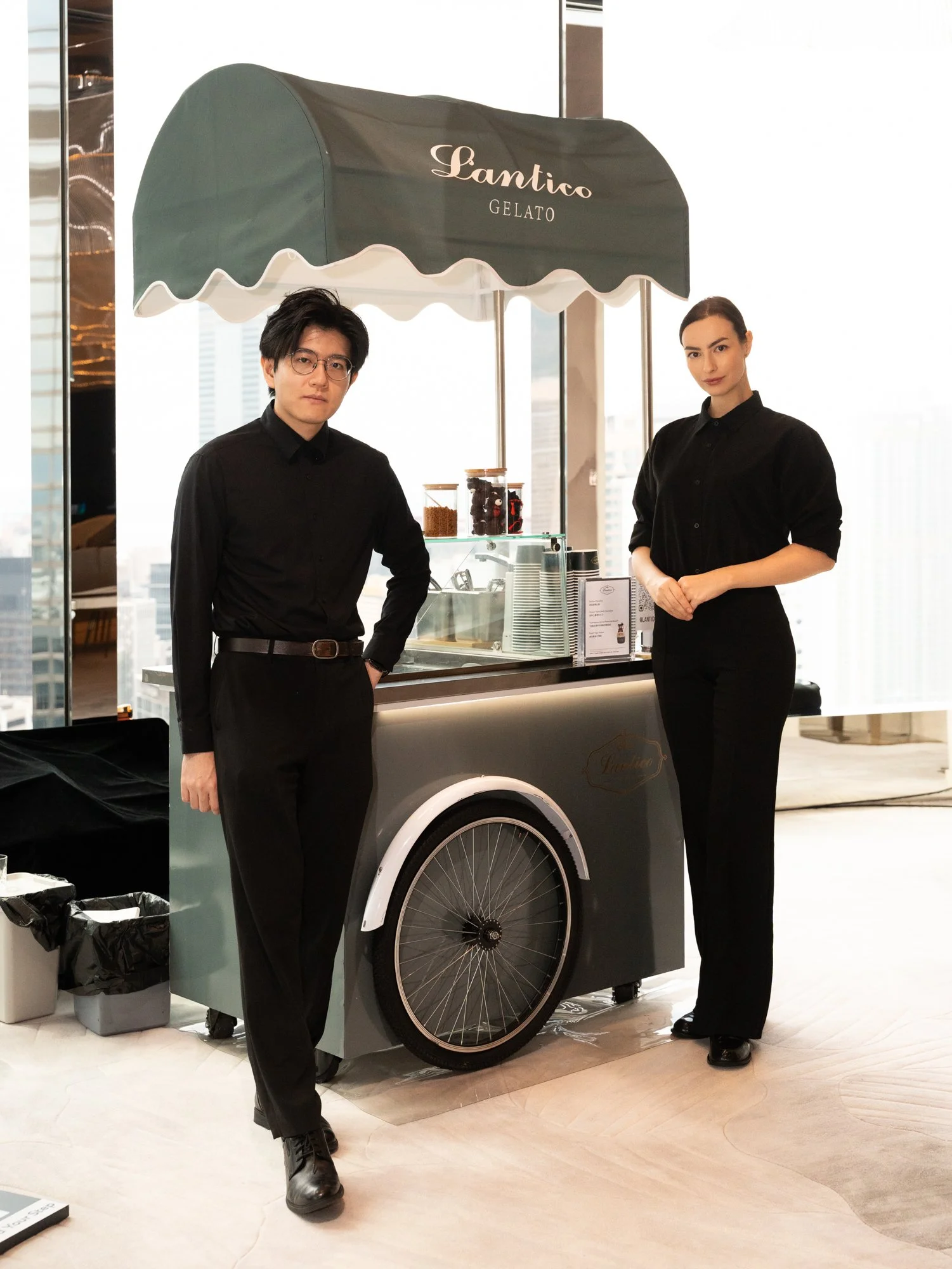 Two staff members stand next to a gelato cart with a green canopy marked 'Lantico Gelato.' The man on the left has black hair, glasses, and is dressed in black clothing. The woman on the right has dark hair pulled back and is also wearing black attir