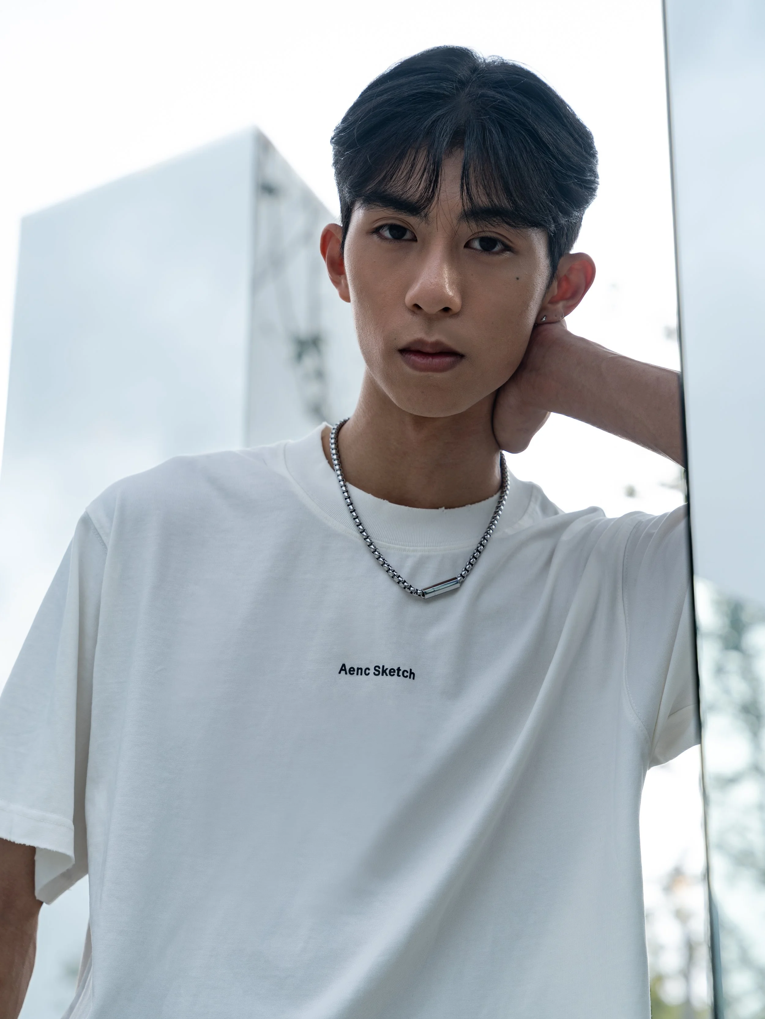 Close-up of a young man with short black hair looking at the camera, wearing a white t-shirt with the text 'Aenc Sketch' and a silver chain necklace, and resting his hand on the back of his neck.