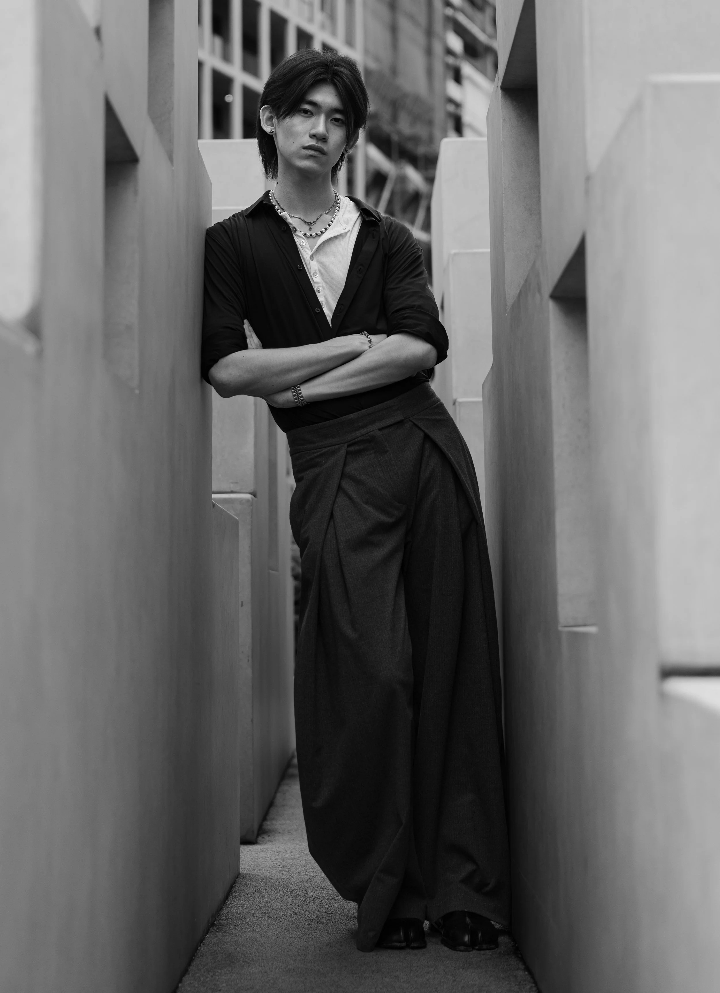 A young man standing in a narrow corridor between large furniture pieces, dressed in dark pants, a light shirt, and a dark open jacket, with crossed arms and looking at the camera.