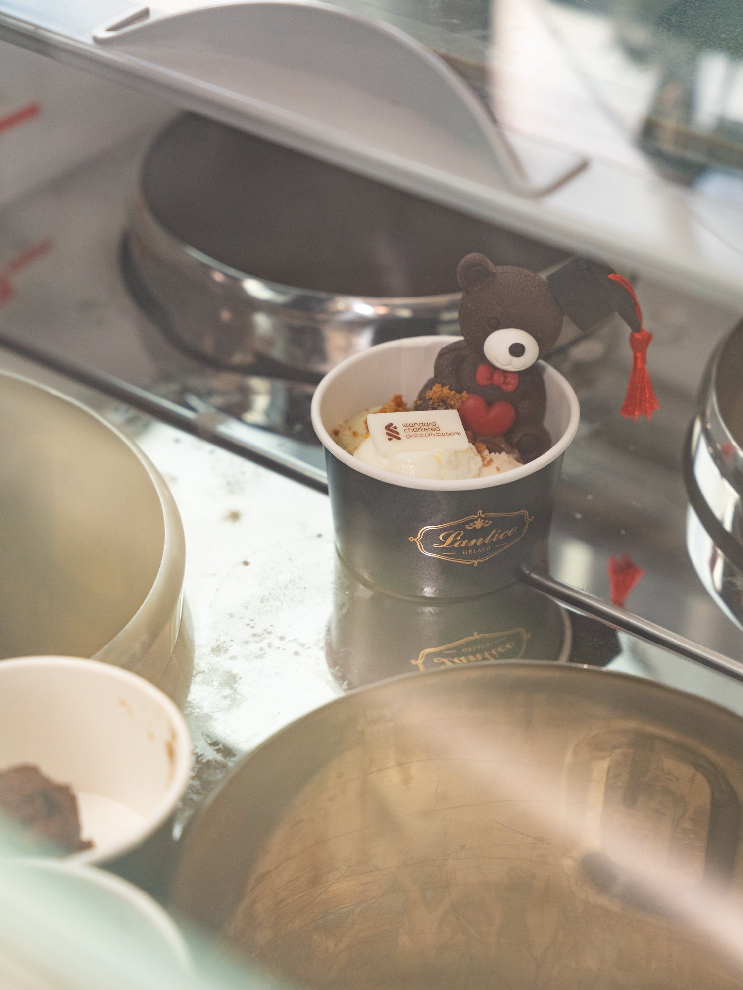 A small black teddy bear figurine with a red bow tie and red heart sitting inside a white cup with a logo, surrounded by empty bowls and a stainless steel countertop.