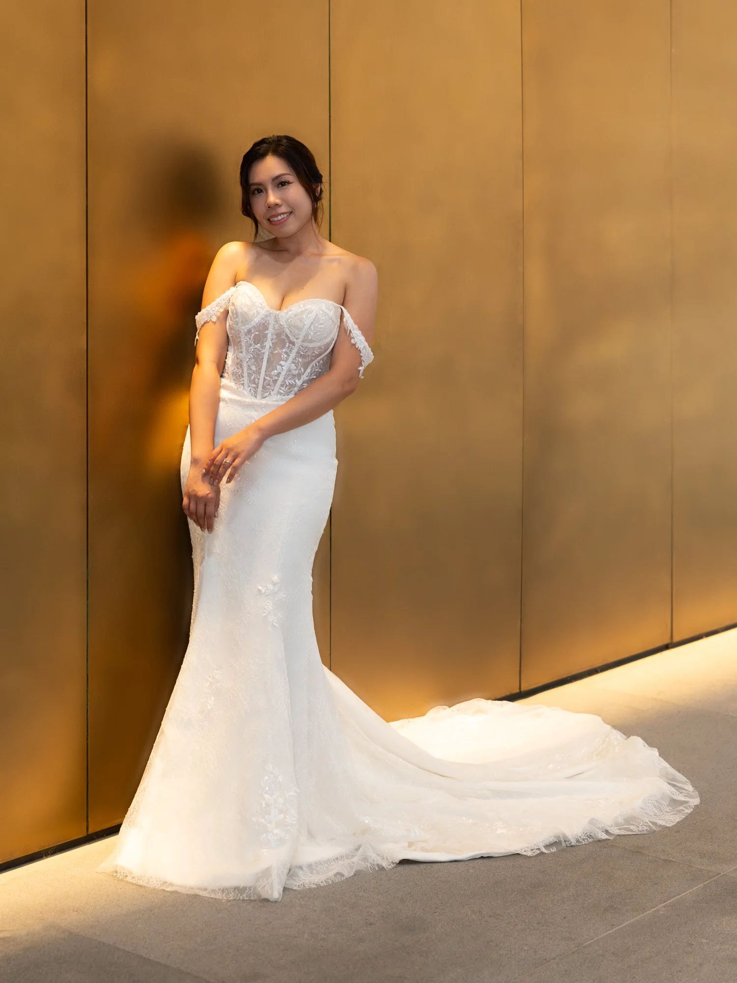 A woman in a white wedding gown stands against a wooden panel wall, smiling at the camera.
