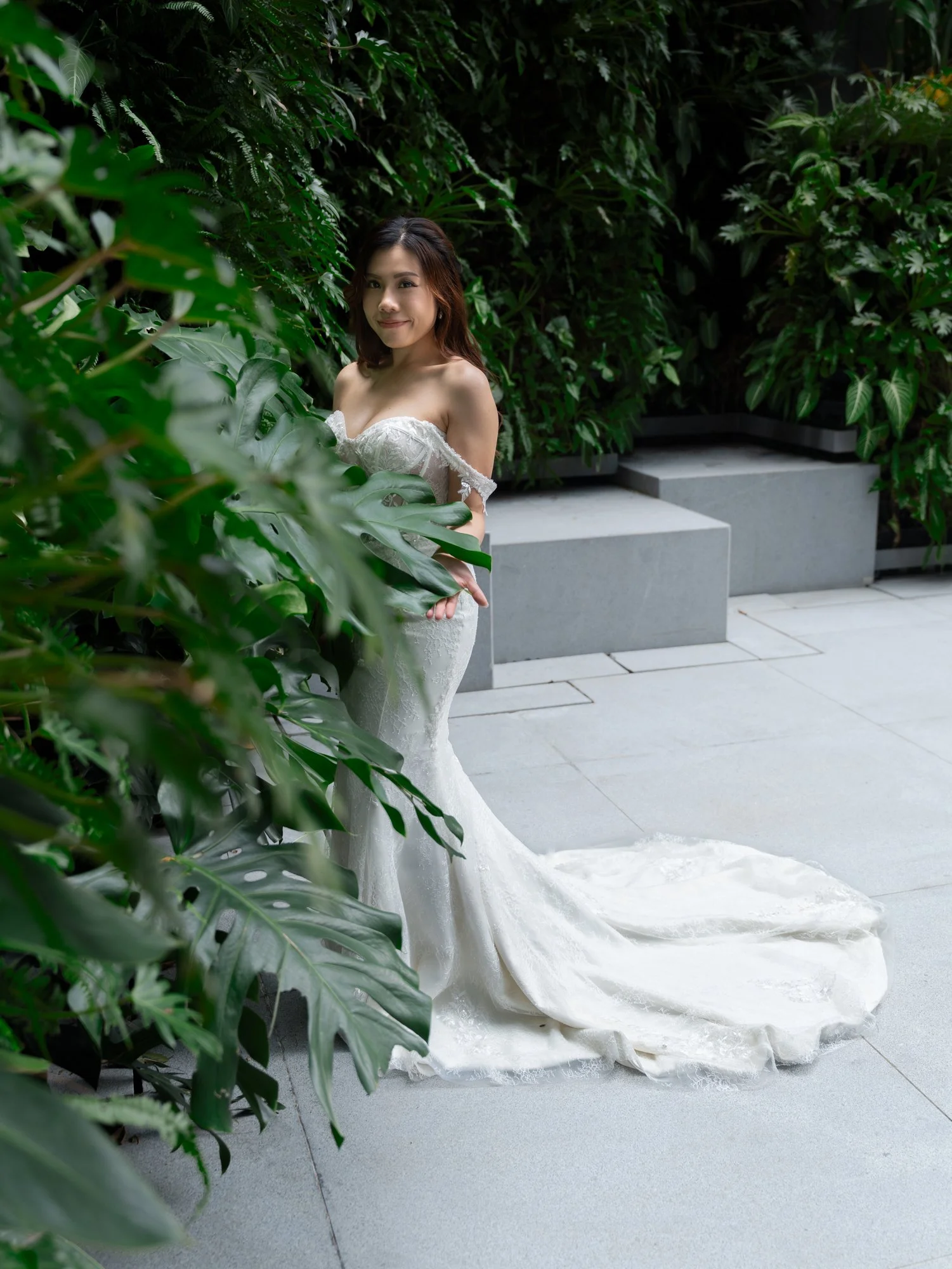 A young woman in a white strapless wedding dress standing behind lush green foliage on a patio with concrete stairs.