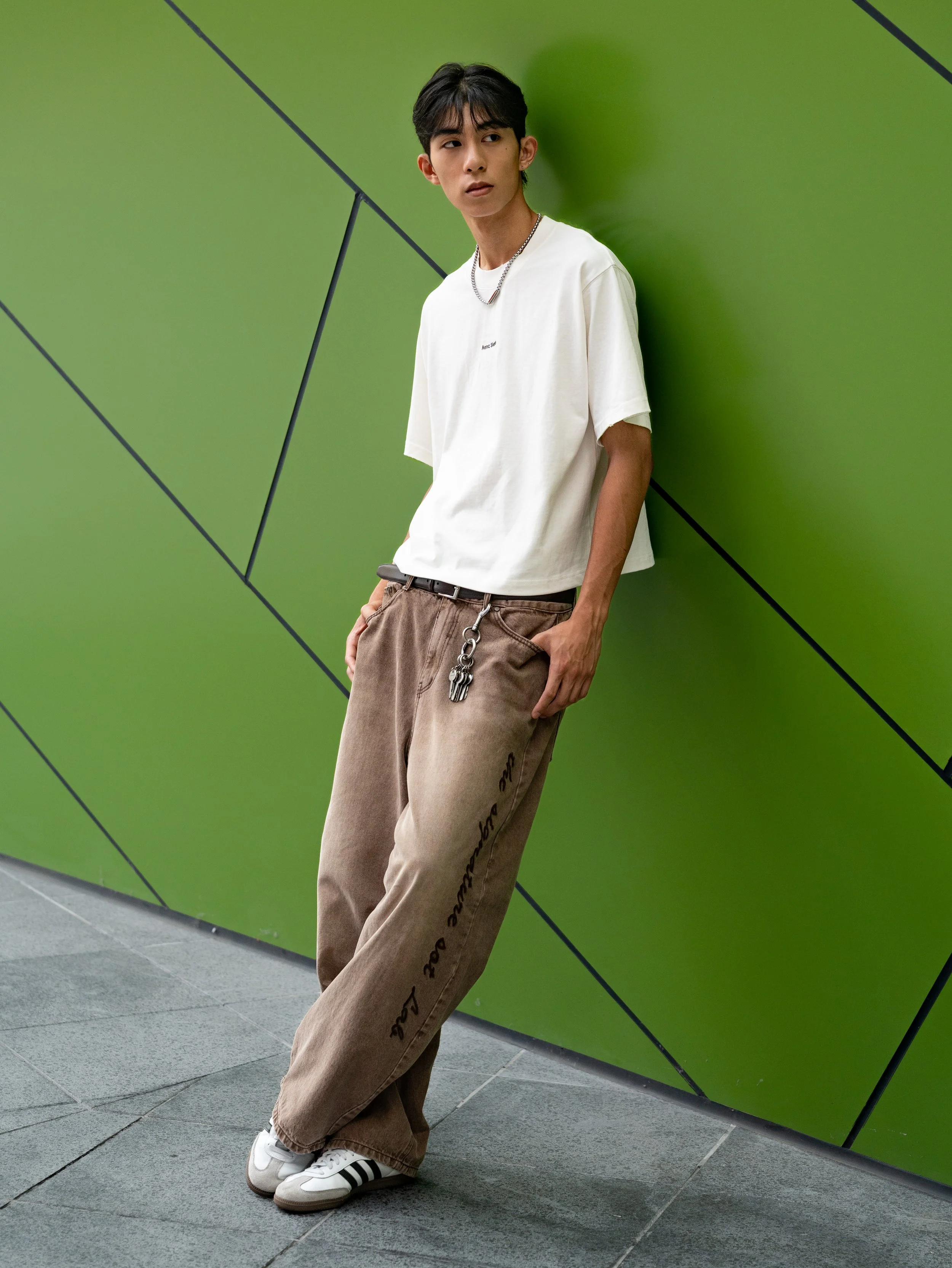 Young man leaning against a green wall with black diagonal lines, wearing a white t-shirt, beige oversized pants with black text, white sneakers with black stripes, and a silver chain necklace.