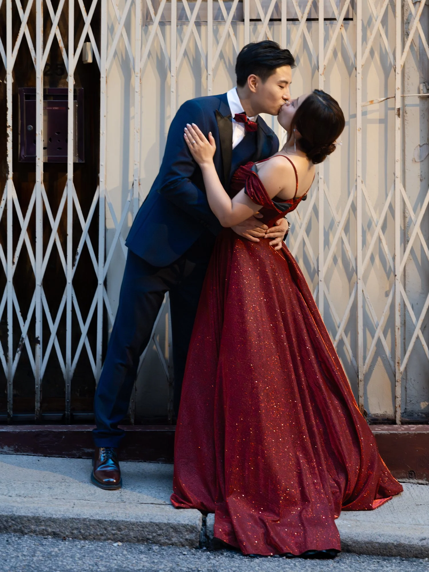 A young couple sharing a kiss outdoors at night. The man is dressed in a dark tuxedo with a red bow tie, and the woman is wearing a strapless, red, glittery evening gown. They are in front of a metal gate with vertical bars.