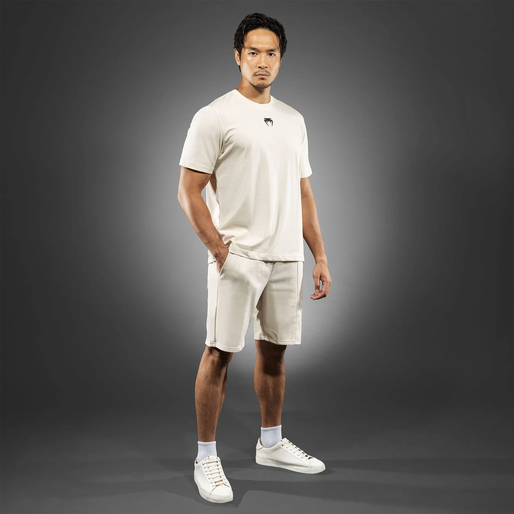 A young man with short black hair standing against a dark gray background, wearing a cream-colored T-shirt with a black graphic, matching shorts, white sneakers, and white socks.