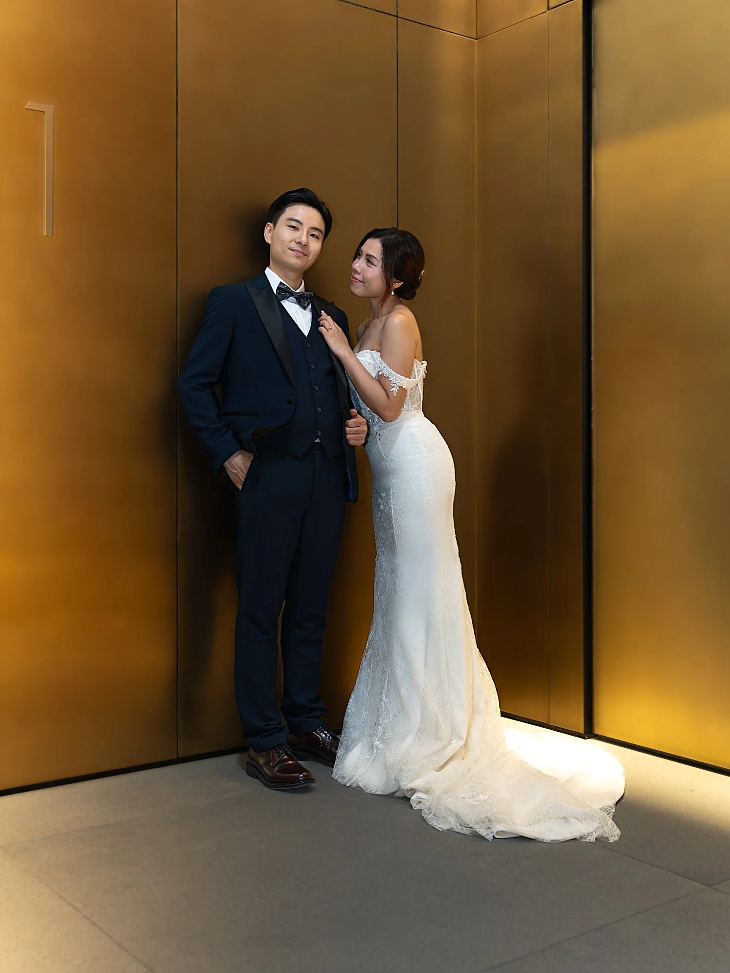 A bride and groom standing in an elevator, with the groom in a navy blue tuxedo and the bride in a white off-the-shoulder wedding gown.