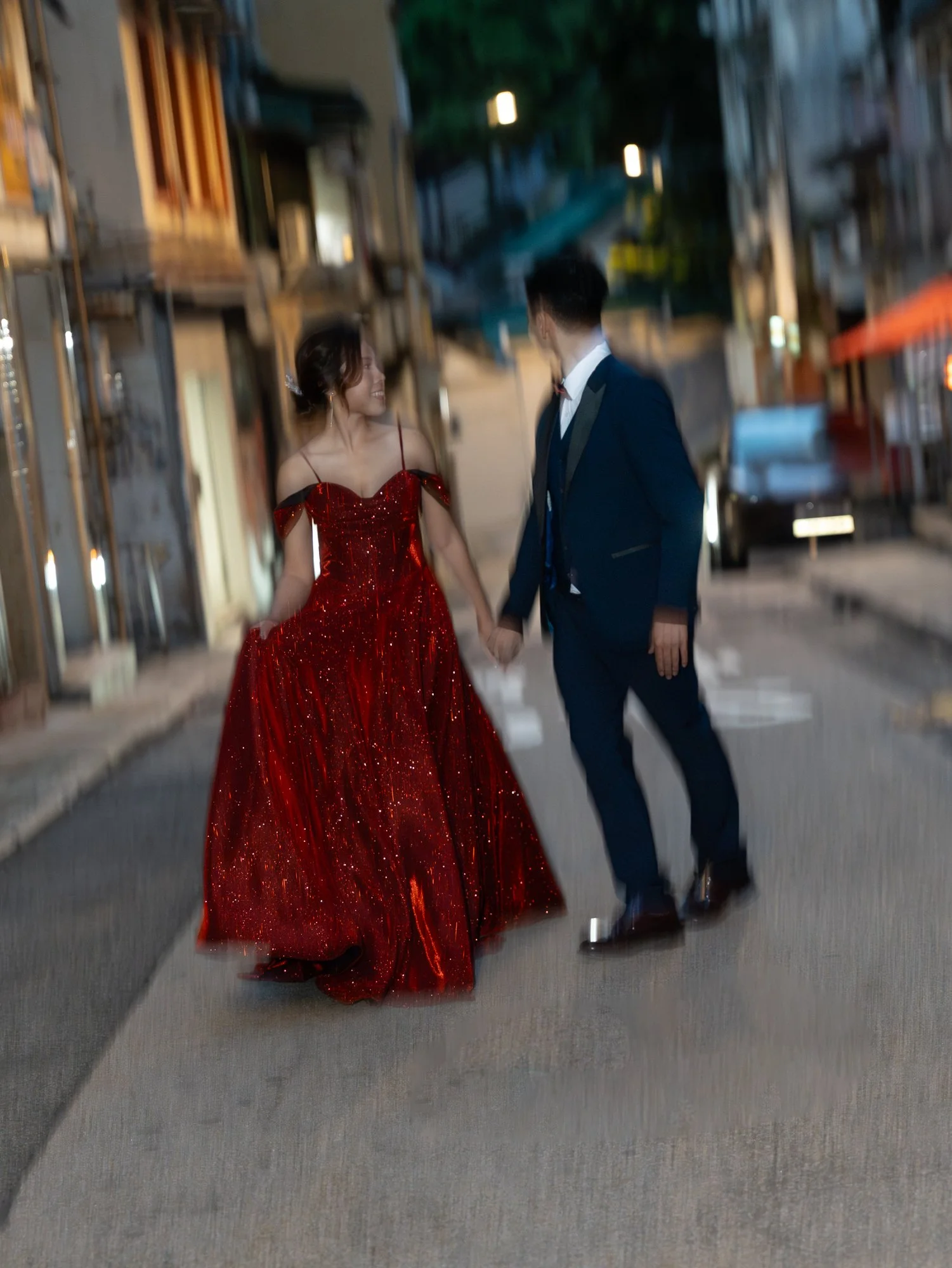 A couple dressed in formal attire holding hands and walking on a gray sidewalk at night, with the woman wearing a red, sparkly off-shoulder gown and the man in a black tuxedo.