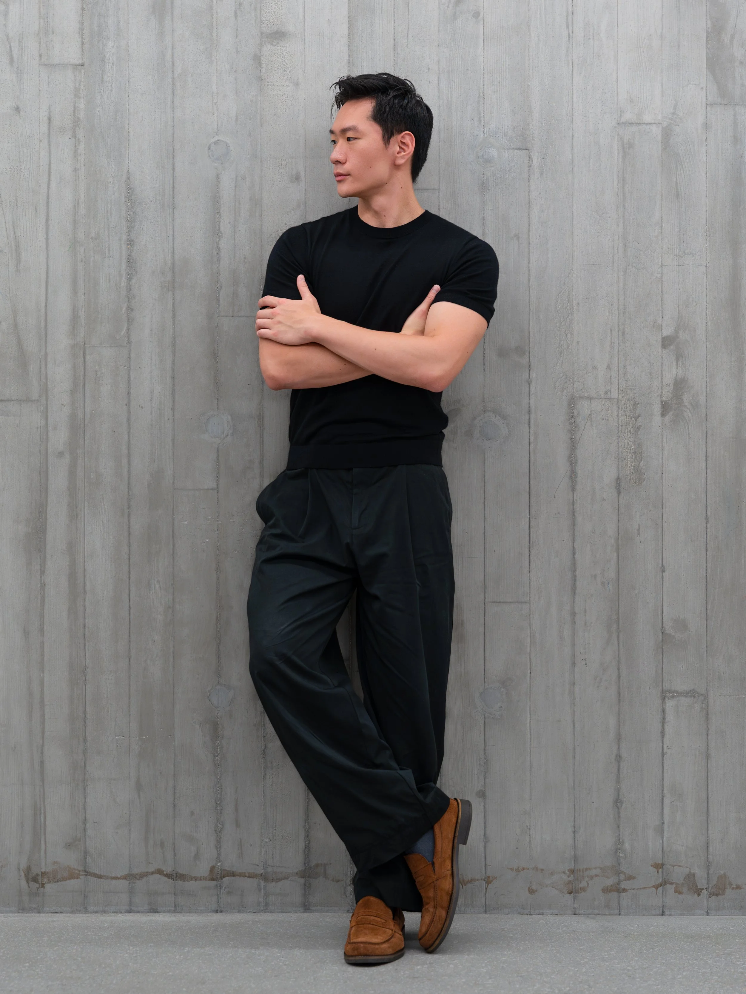A young man with dark hair in a casual pose stand against a gray concrete wall, crossing his arms and looking to the left. He wears a black t-shirt, black pants, and brown shoes.