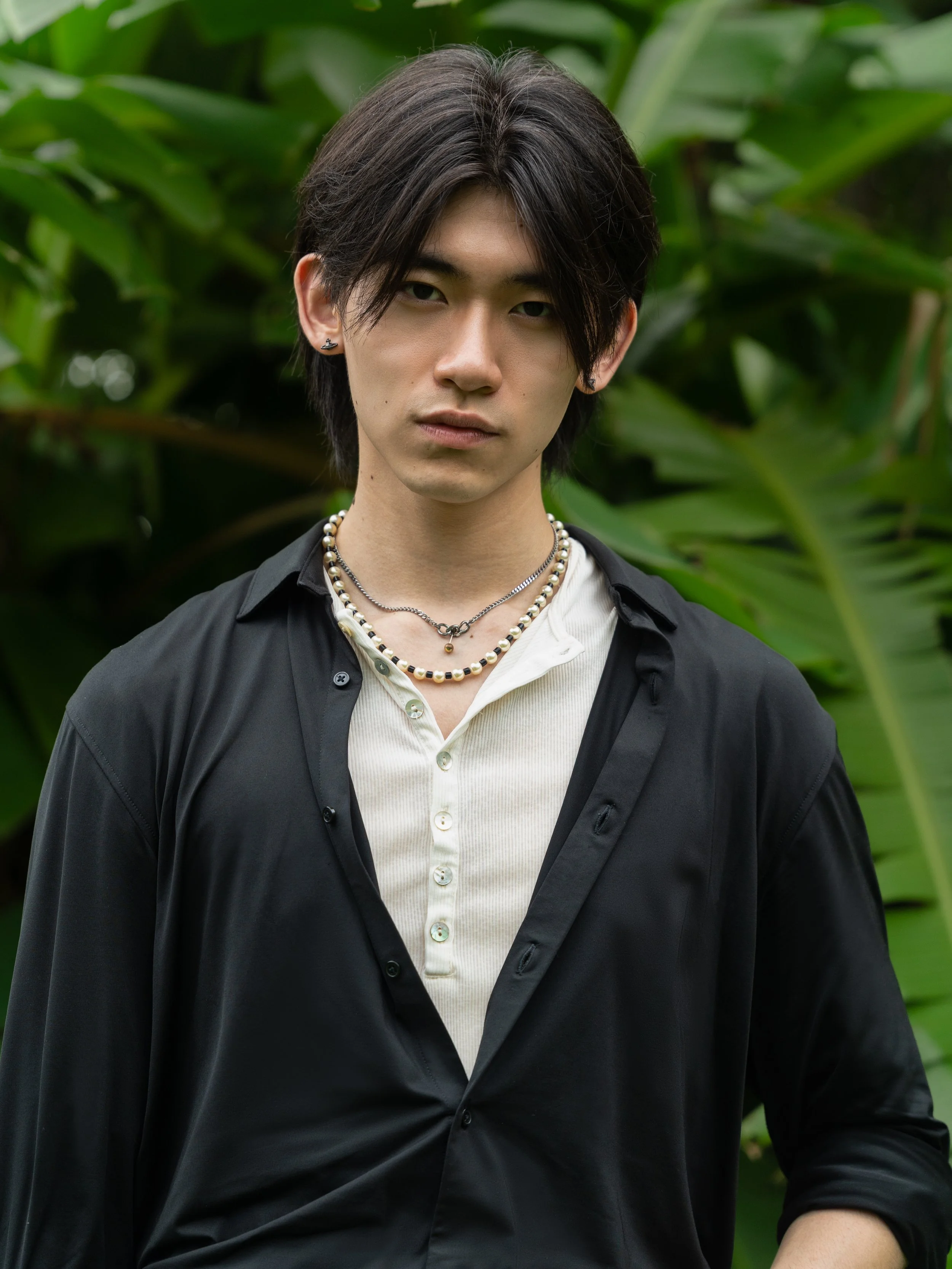 A young man with medium-length dark hair, wearing multiple necklaces, earrings, and a black shirt with an open collar, standing outdoors in front of lush green plants.