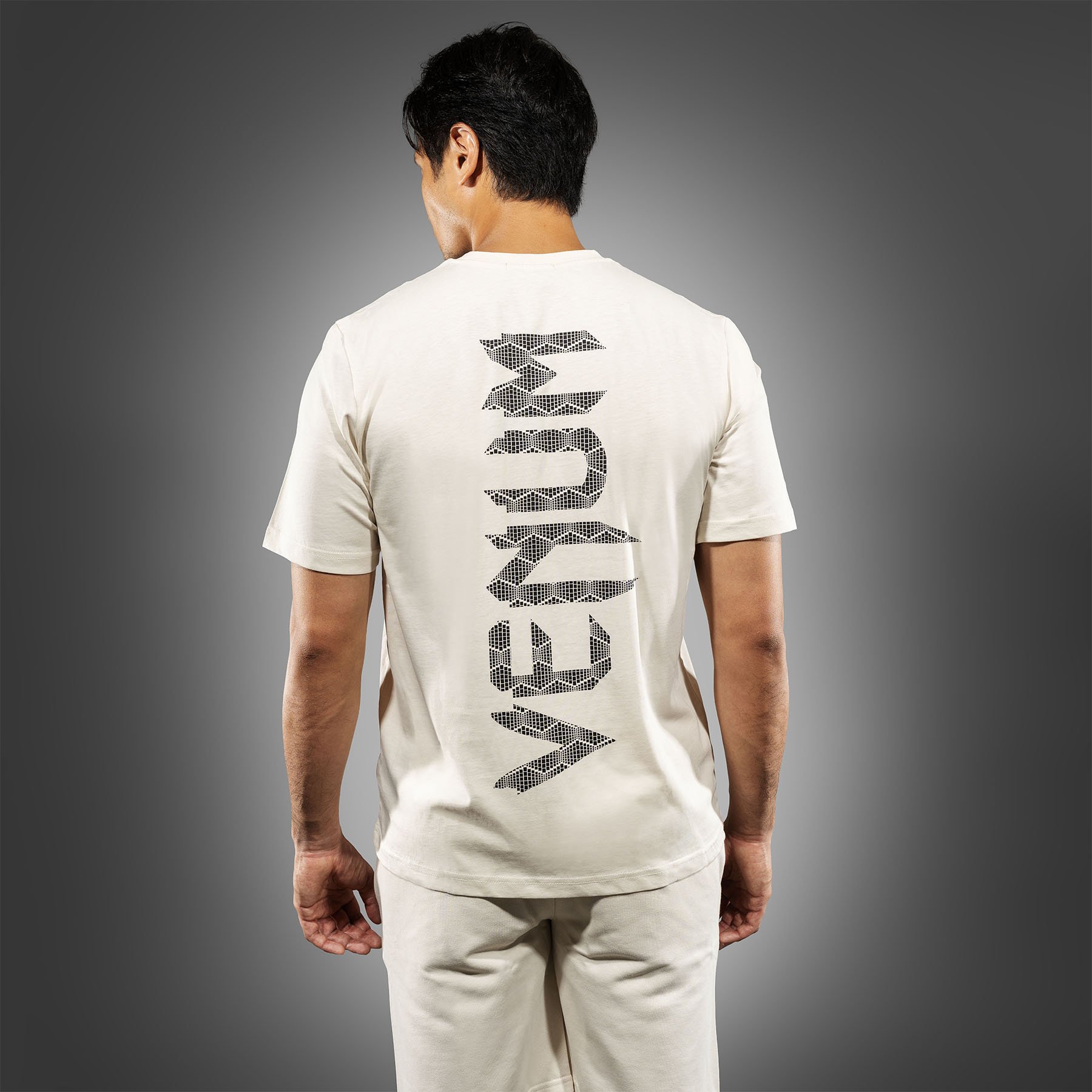 Back view of a man with dark hair wearing a white T-shirt and beige pants against a gray background. The T-shirt has the word "WALK" printed vertically in large, pixelated black letters on the back.