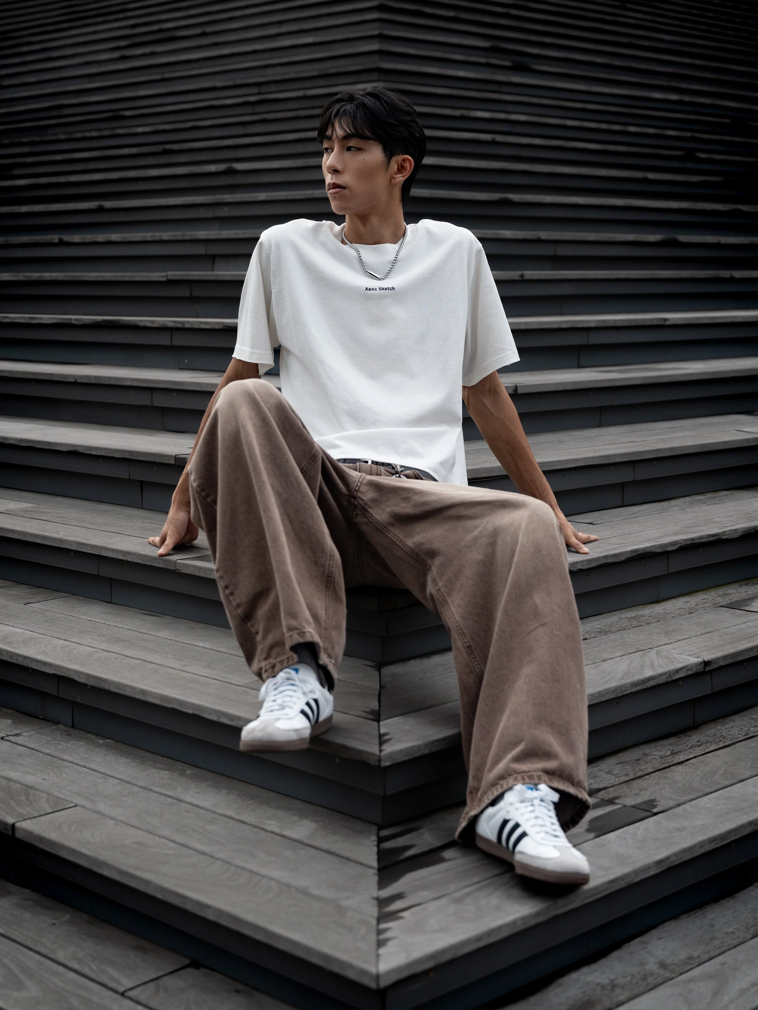 A young man with short dark hair sitting on a set of outdoor wooden stairs wearing a white t-shirt, beige pants, and white sneakers with black stripes, looking to the side.