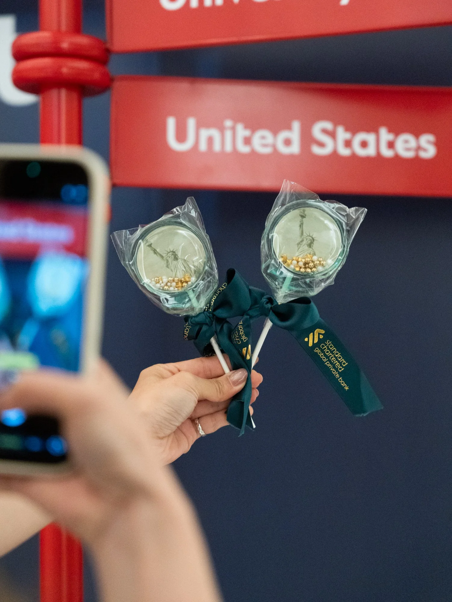 Person holding two lollipops with small beads in clear plastic wrappers, standing in front of a sign that reads 'United States', with part of a phone or camera visible in the foreground.